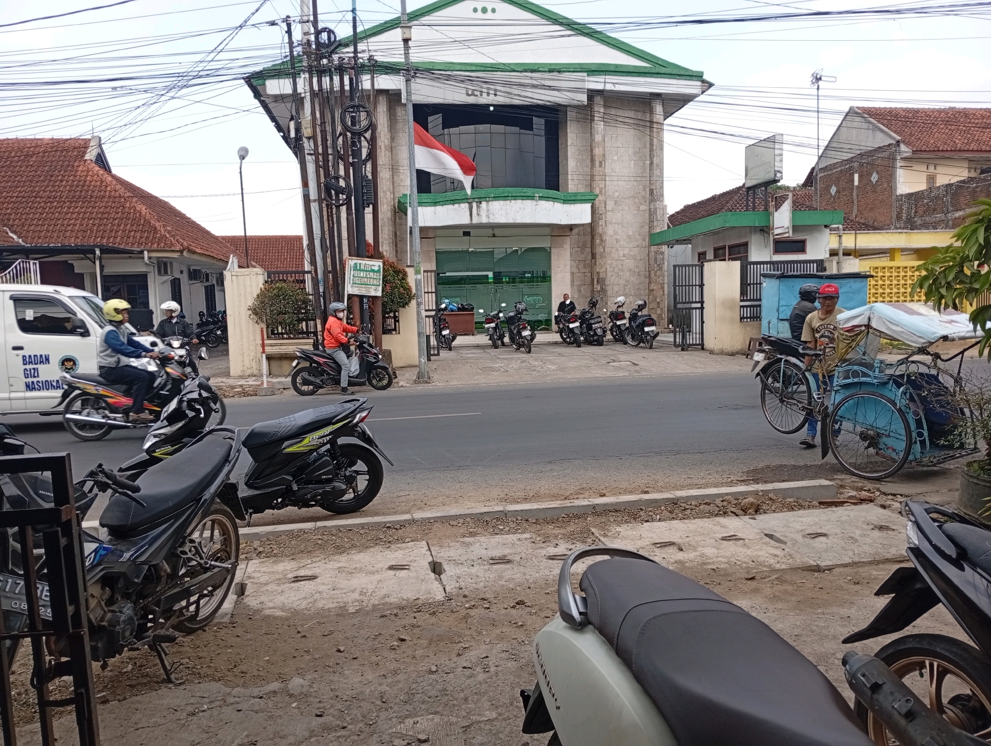 This is a daytime, eye-level, medium-wide shot of a street scene in Tasikmalaya, Indonesia, near a building identified by a sign as "PUSKESMAS SIGEUREUNG." The building, which appears to be a public health center, is light-colored with a green roof and features a large Indonesian flag displayed prominently. The street in front of the building is paved, with a sidewalk on the right side.
Several motorcycles are parked on the sidewalk and in the street. On the right side of the street, a man is operating a blue three-wheeled bicycle cart. Further down the street, a white van with text identifying it as "BADAN GIZI NASIONAL" (National Nutrition Agency) is visible.
Several people are present, mostly on or around motorcycles. One person is riding a motorcycle in the foreground. Another person is seen near the three-wheeled cart. A few individuals are standing near the entrance of the PUSKESMAS SIGEUREUNG building. The sky is overcast. The environment suggests a busy urban setting.