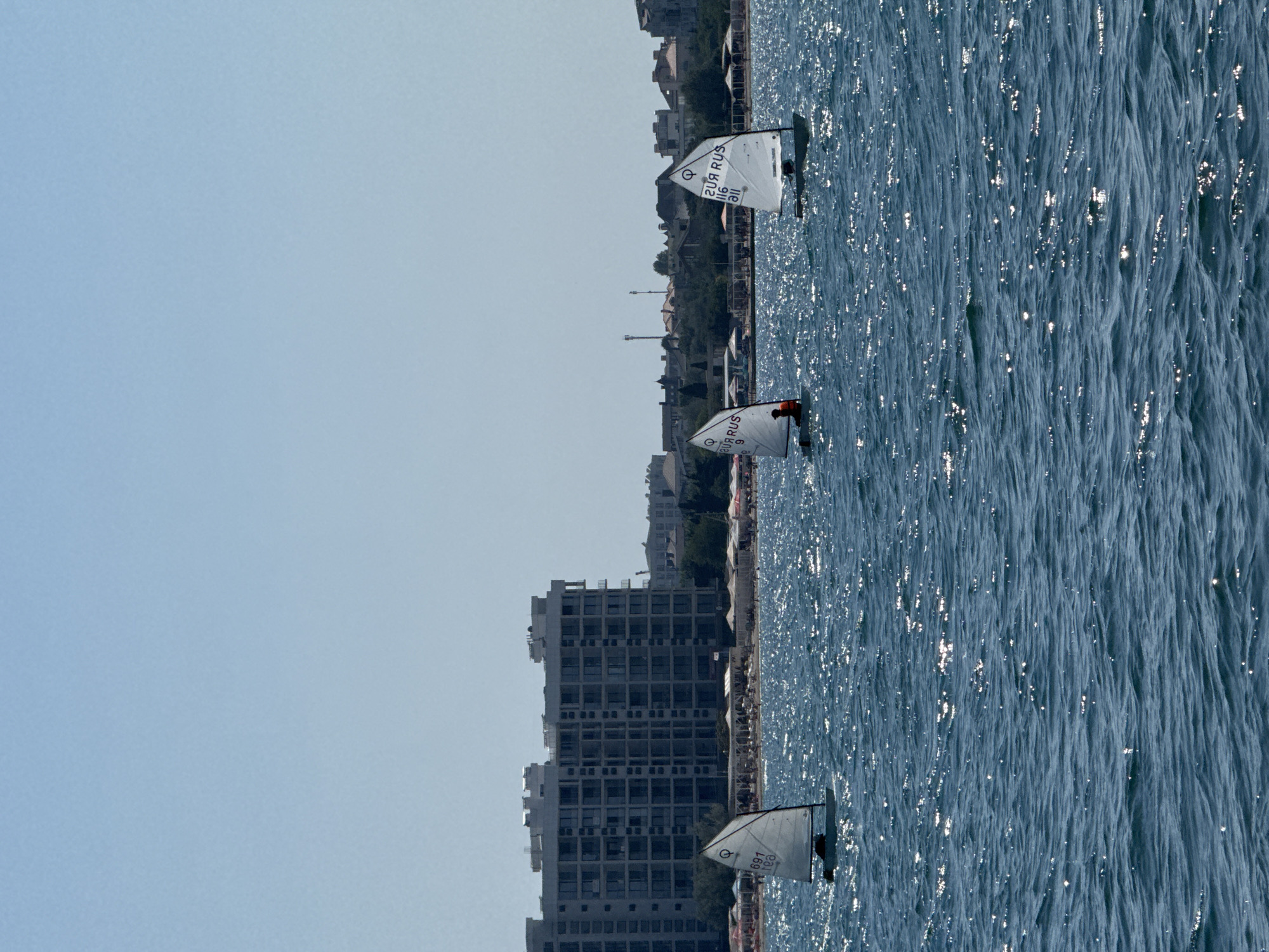 In Sevastopol, Ukraine, on a sunny day, three small sailboats are navigating the dark blue, choppy waters of the sea. The sailboats are close to each other, with one in the foreground and two further back to the right. Each sailboat has a white sail with markings that appear to be in Cyrillic. The sailboat on the left has the number "691" and the letters "rea" visible on its sail. The middle sailboat shows "20R RUS 116" and the last one, on the right, displays "20R RUS 116 all". The water is illuminated by bright sunlight, creating a shimmering effect across its surface. In the background, a large, multi-story modern building with numerous windows stands prominently. Behind the building, a shoreline is visible with trees and smaller structures, suggesting a coastal city. The sky is a clear, pale blue with no clouds.