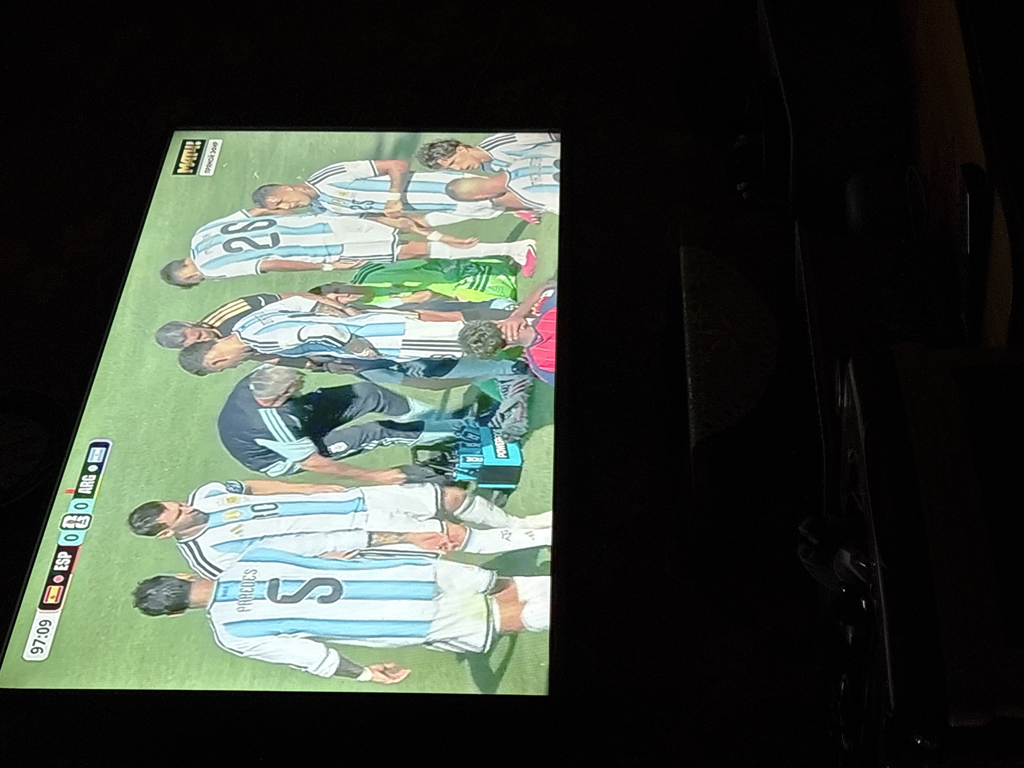 A television screen displays a soccer match between Spain (ESP) and Argentina (ARG). The score is 0-0 at 97:09. Several players in Argentina's white and light blue striped jerseys are visible, some with their backs to the camera. One player, number 5, has "PAREDES" printed on his jersey. Another player, number 10, is also visible. A player in a dark uniform is bent over, possibly tending to an injury. A bright blue medical bag with the word "POWERADE" is on the grass. A green soccer goalie uniform is also visible on the field. The background is a green soccer field. The television is dark and shows no indication of being in a specific city or location withinВетлужский, Russia.