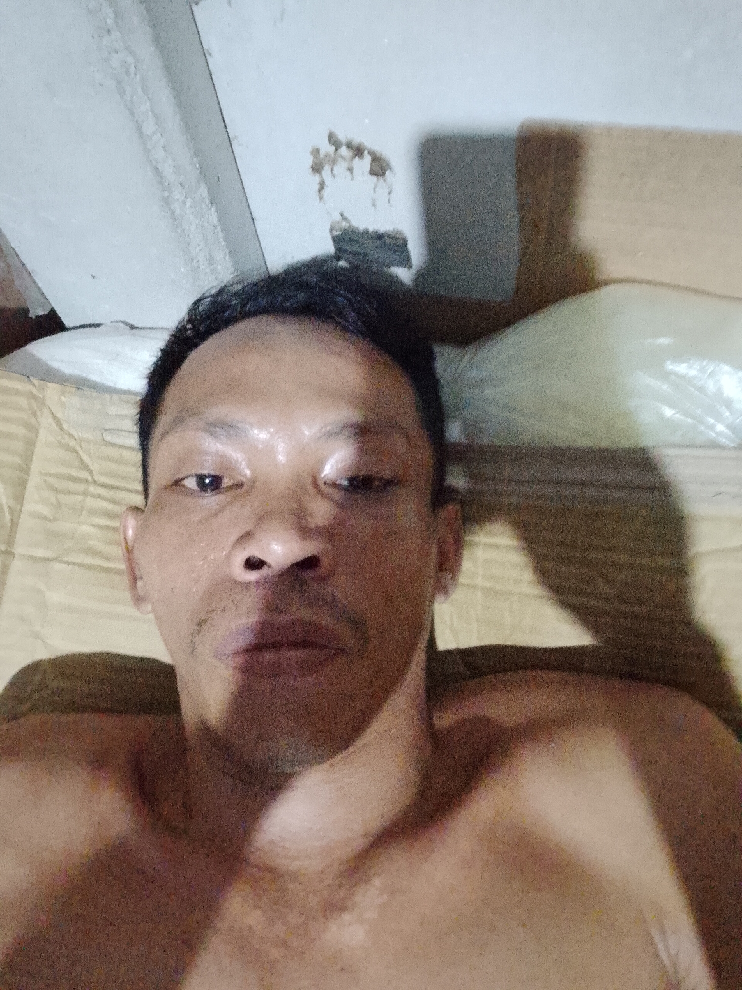 The image depicts a shirtless individual, possibly male, with dark hair, lying on a yellow patterned surface resembling a bed or mattress. The person is facing the camera, with a neutral expression. The skin appears slightly damp or sweaty.
The background is composed of a white wall with what appears to be water damage or staining near the top left corner. A cardboard box or similar rectangular object is partially visible behind the person's head. The location is implied to be Bukit Ketok, Indonesia, based on the prompt. No other specific details about the location are discernible from the image.