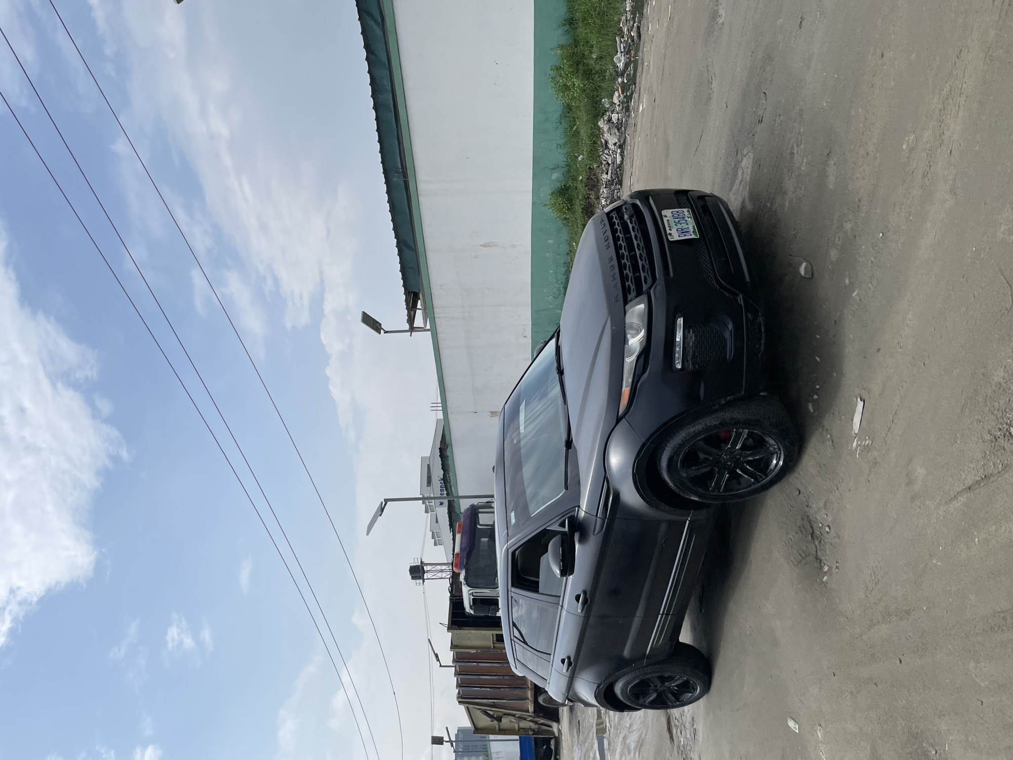 A black Range Rover Evoque is parked on a dirt road in Igboefon, Nigeria. The car is facing left and is viewed from the front three-quarters. It has black rims and tinted windows. A license plate is visible on the front of the car, displaying "ABUJA" and "BWR 354BB". In the background, there is a white wall with a green stripe and some greenery. To the left of the car, a large refuse truck is parked. The sky is blue with scattered clouds. The ground is uneven and appears to be a mix of dirt and gravel with some scattered debris. Overhead, power lines are visible.