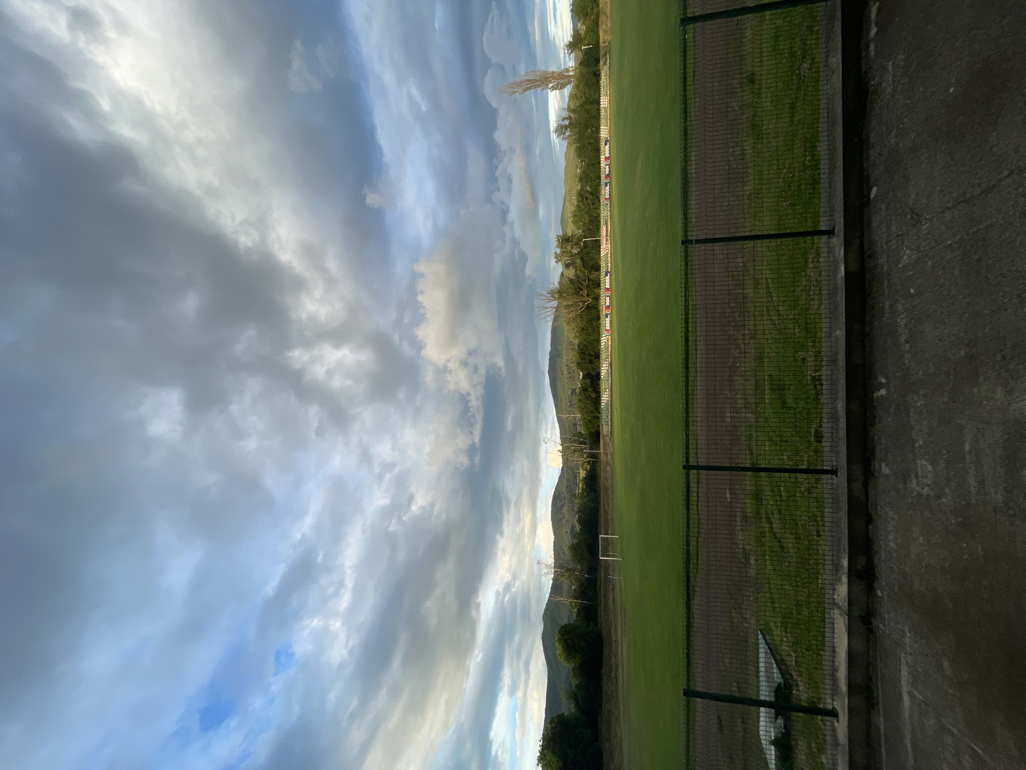 The image, originally captured in portrait orientation, displays a wide landscape view of a sports facility under a cloudy sky.
In the immediate foreground, to the right, is a section of concrete or paved ground. This area is bordered by a dark, mesh fence that extends horizontally across the lower right part of the frame, separating it from a vibrant green sports field. The field, likely a football or soccer pitch, occupies the central midground. A small white goalpost is visible on the left side of the field. Along the far sideline of the field, a series of advertising banners are prominently displayed. Beyond the field and banners, scattered trees punctuate the landscape. The background features a range of rolling hills or mountains, covered with trees and foliage, extending into the distance. Dominating the upper portion of the image is a vast sky, filled with an array of dramatic, textured clouds. Patches of brighter, illuminated clouds contrast with darker, more volumetric ones, with occasional glimpses of blue sky peeking through.
No specific activity or event is taking place, and no individuals are visible, suggesting a calm and unoccupied scene.
The time of day appears to be late afternoon or early evening, evidenced by the soft, warm light that highlights the cloud formations and illuminates parts of the landscape. The weather is partly cloudy, showcasing a mix of bright and shaded areas across the sky. The overall atmosphere conveys tranquility and spaciousness, typical of a rural or semi-rural setting. The mountainous backdrop is consistent with the provided location context of Ihtiman, Bulgaria.
The advertising banners on the far side of the sports field clearly display the repeating text "888BET".