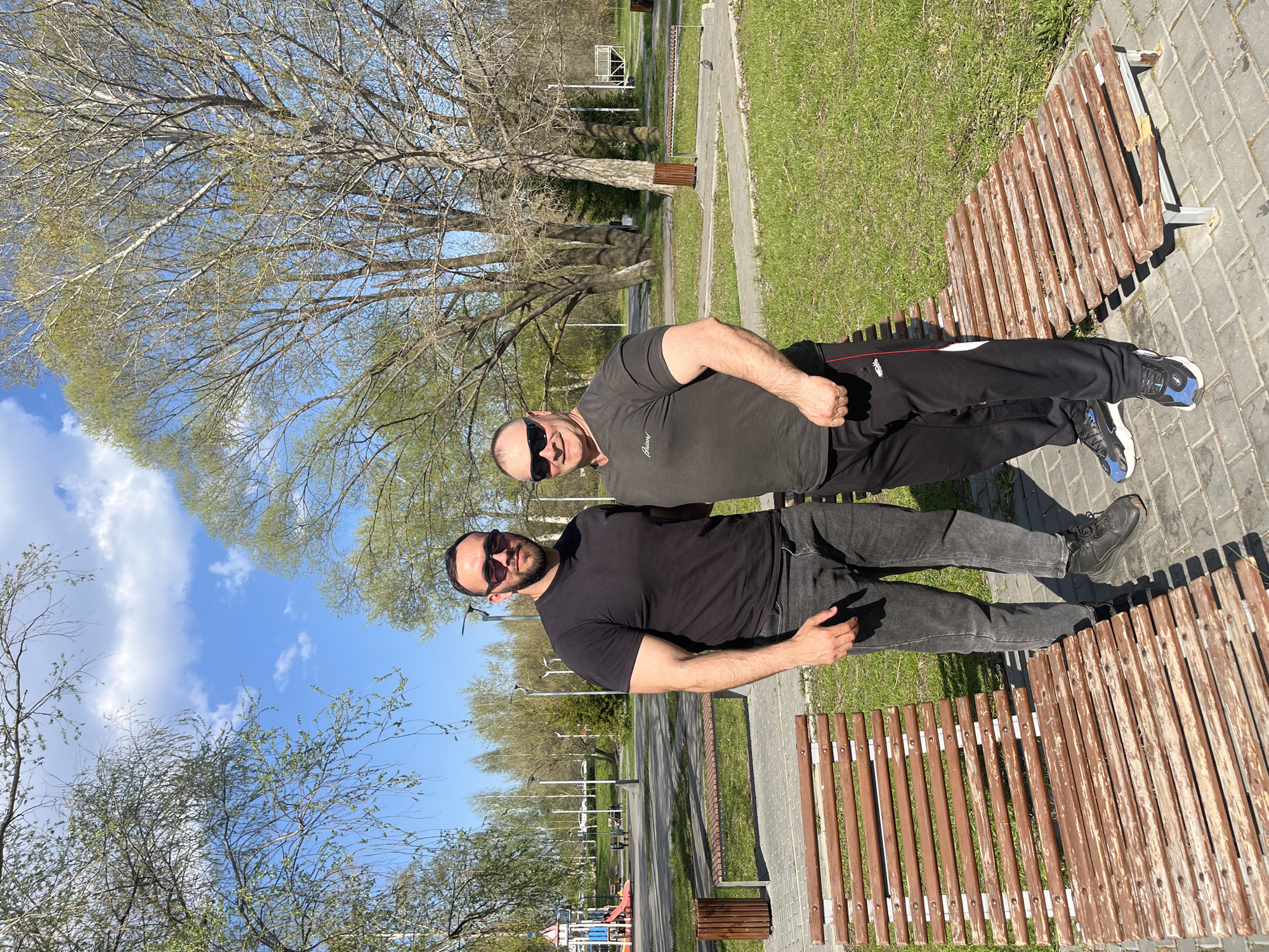 Two men wearing sunglasses are sitting on a wooden bench in a park. The man closer to the camera wears a black t-shirt and dark gray jeans with black shoes. The other man wears a dark gray t-shirt and black tracksuit pants with blue and black shoes. The bench is located on a brick walkway in a grassy area with trees and bushes. In the background, there is a road with lampposts and a children's playground. Based on the location, the image may be taken in a park in Ulyanovsk, Russia.