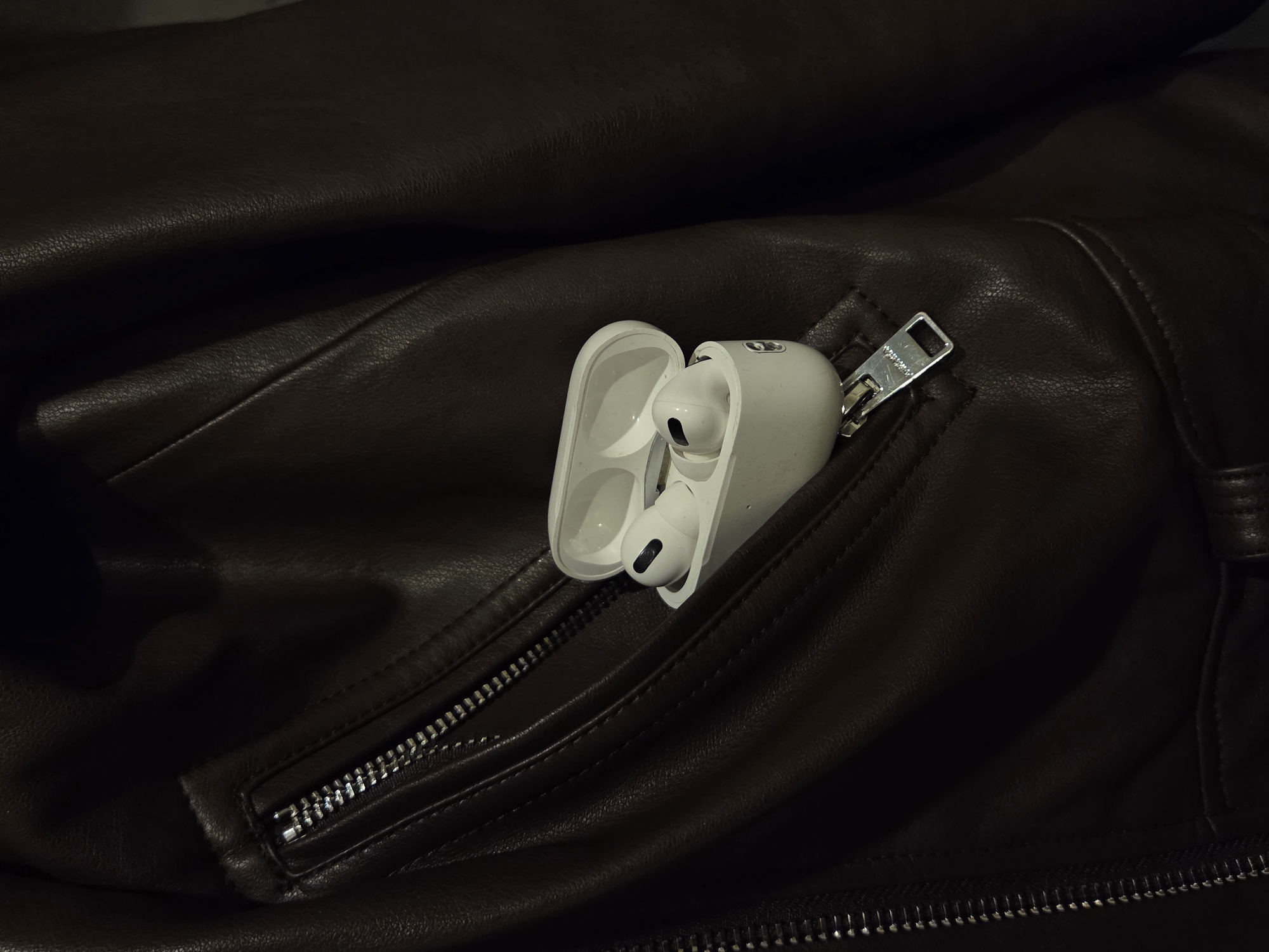 A white, open, wireless-earphone charging case containing two white wireless earphones is in the open zippered pocket of a brown leather jacket. The zipper pull is silver. The location is Unknown, Unknown.
