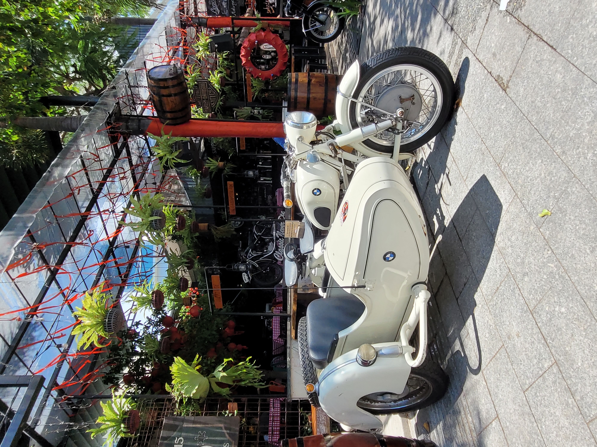 The image captures a light beige vintage BMW motorcycle with a sidecar parked on a gray-tiled pedestrian walkway. A modern-style canopy structure spans above the motorcycle, adorned with hanging plants, barrels, and red ribbons, creating a decorative urban space. In the background, more motorcycles and outdoor seating can be seen. The setting appears to be a café or restaurant area within a city.