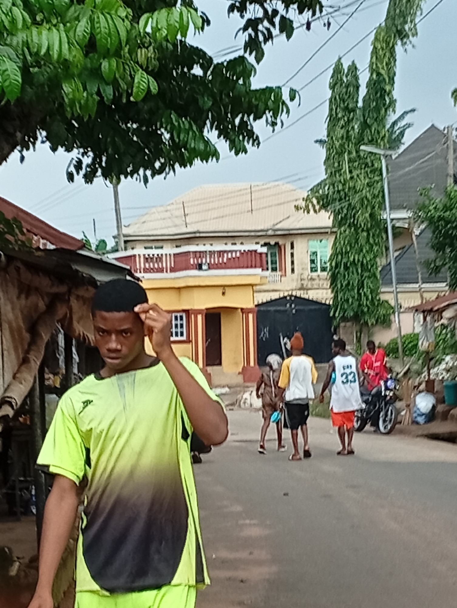The image depicts a street scene in Nsugpe, Nigeria. In the foreground, a young man wearing a neon yellow and black t-shirt is looking towards the camera and touching his head. In the background, several people are walking on the street. One person is wearing a white tank top with the number "33" and orange shorts, another is in a red shirt, and two others are in more traditional attire. A motorcycle is parked on the side of the road. The environment features buildings with a mix of modern and traditional architecture, some with visible metal roofs and decorative elements. Green trees and power lines are also present in the scene. The overall impression is a snapshot of everyday life on a street in Nsugpe.