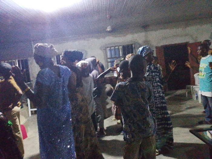 A group of people gather indoors. Some appear to be dancing or moving rhythmically. Individuals wear patterned clothing; one person is in a blue dress. A man in a blue t-shirt stands near a doorway. The lighting is dim, and the room has a simple, utilitarian appearance with chairs visible near the walls. The atmosphere suggests a community gathering, possibly with a spiritual or celebratory context.