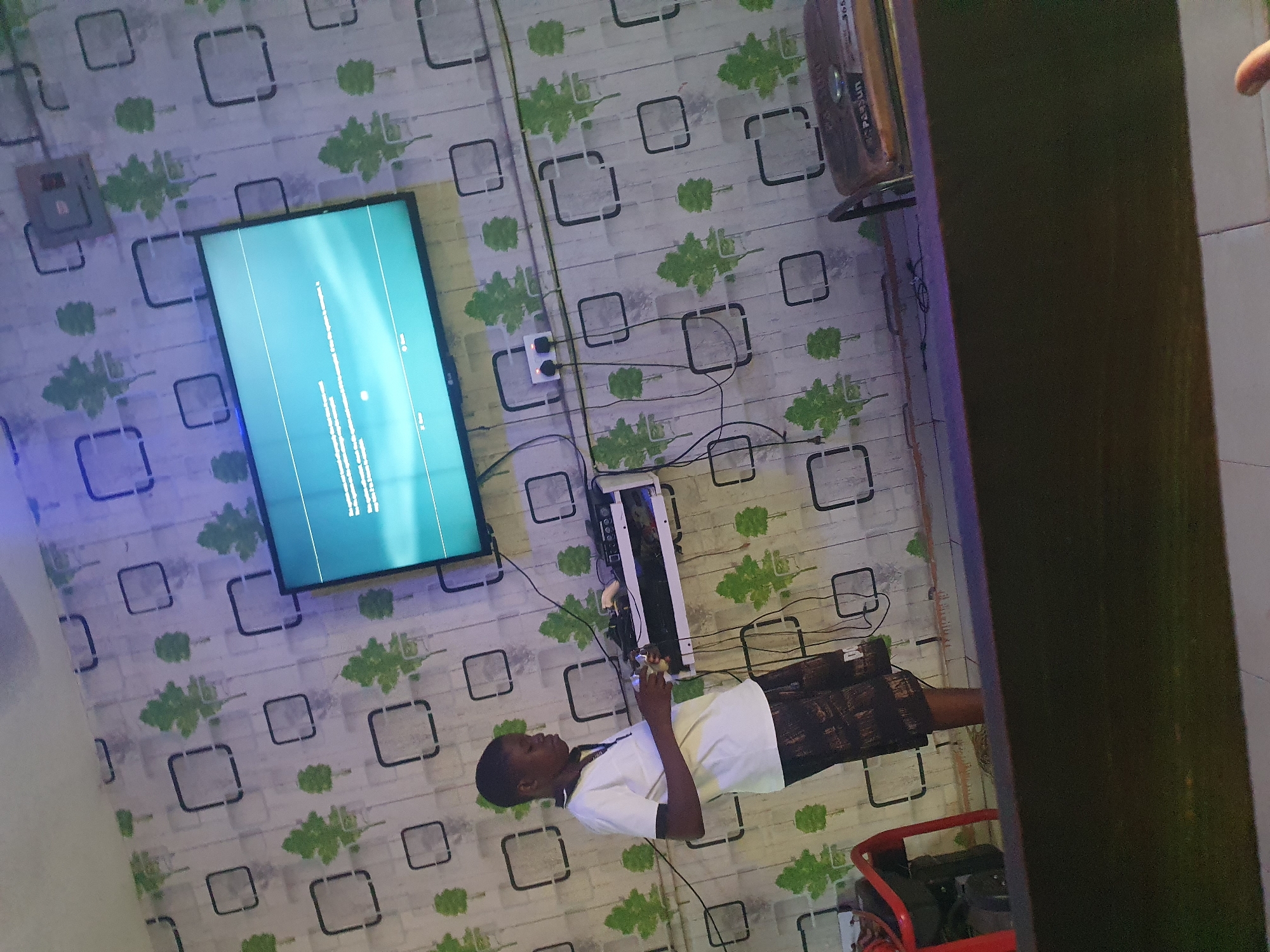 A young Black male, wearing a white t-shirt and patterned shorts, stands in front of a wall adorned with green tree-shaped wallpaper. He is holding a video game controller and looking towards a television screen mounted on the wall. The TV displays a blue screen with text. Below the TV, a white shelf holds electronic equipment and wires. A red chair is visible to the left of the frame, and a brown wooden object is to the right. The scene appears to be indoors, possibly in a living room or entertainment area.