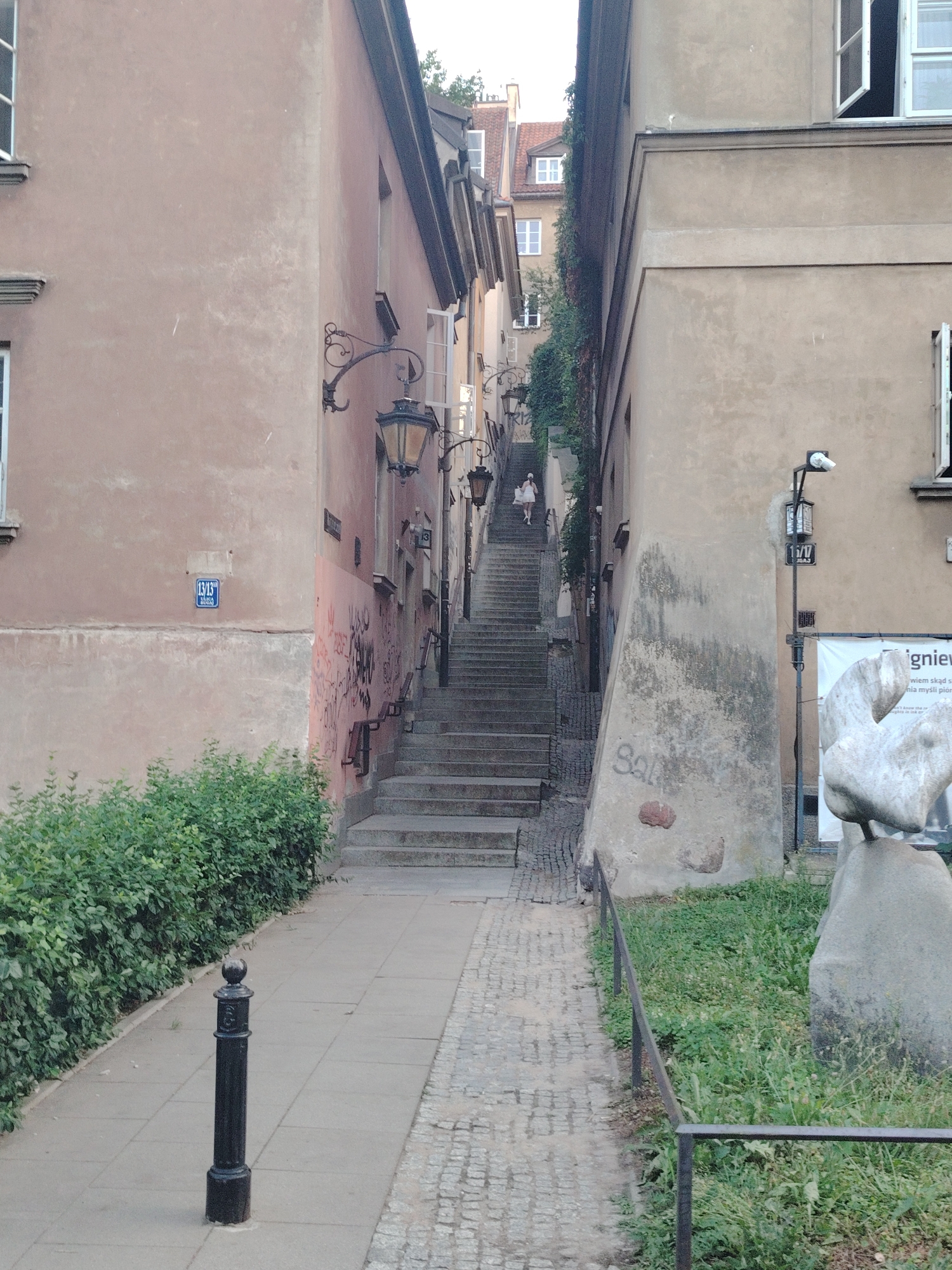 This media file depicts a narrow cobblestone street in Warsaw, Poland, flanked by tall, aged buildings. The architecture suggests an older European city, with stucco walls painted in muted tones of beige and pink. A steep, stone staircase dominates the center of the image, leading upwards between the buildings. At the top of the stairs, a person dressed in white can be seen walking down. Traditional-style street lamps with ornate metalwork are mounted on the walls of the buildings, illuminating the pathway.
The scene appears to be a quiet, possibly historic, alleyway. The lighting suggests it is daytime, with overcast or diffused sunlight, as there are no harsh shadows. The overall mood is serene and somewhat nostalgic, evoking a sense of timelessness. Greenery, including bushes and ivy, grows along the sides of the buildings and in small patches of ground, adding a touch of nature to the urban environment.
Visible text includes house numbers on blue plaques, such as "13/13" and "1/17". There is also graffiti on the pink wall of the building on the left. A large banner with text that appears to be in Polish is visible on the building to the right of the stairs, featuring the name "igniew". A stone sculpture of what looks like a bird or an abstract form is situated to the right of the banner. A black, decorative bollard stands on the tiled sidewalk to the left.