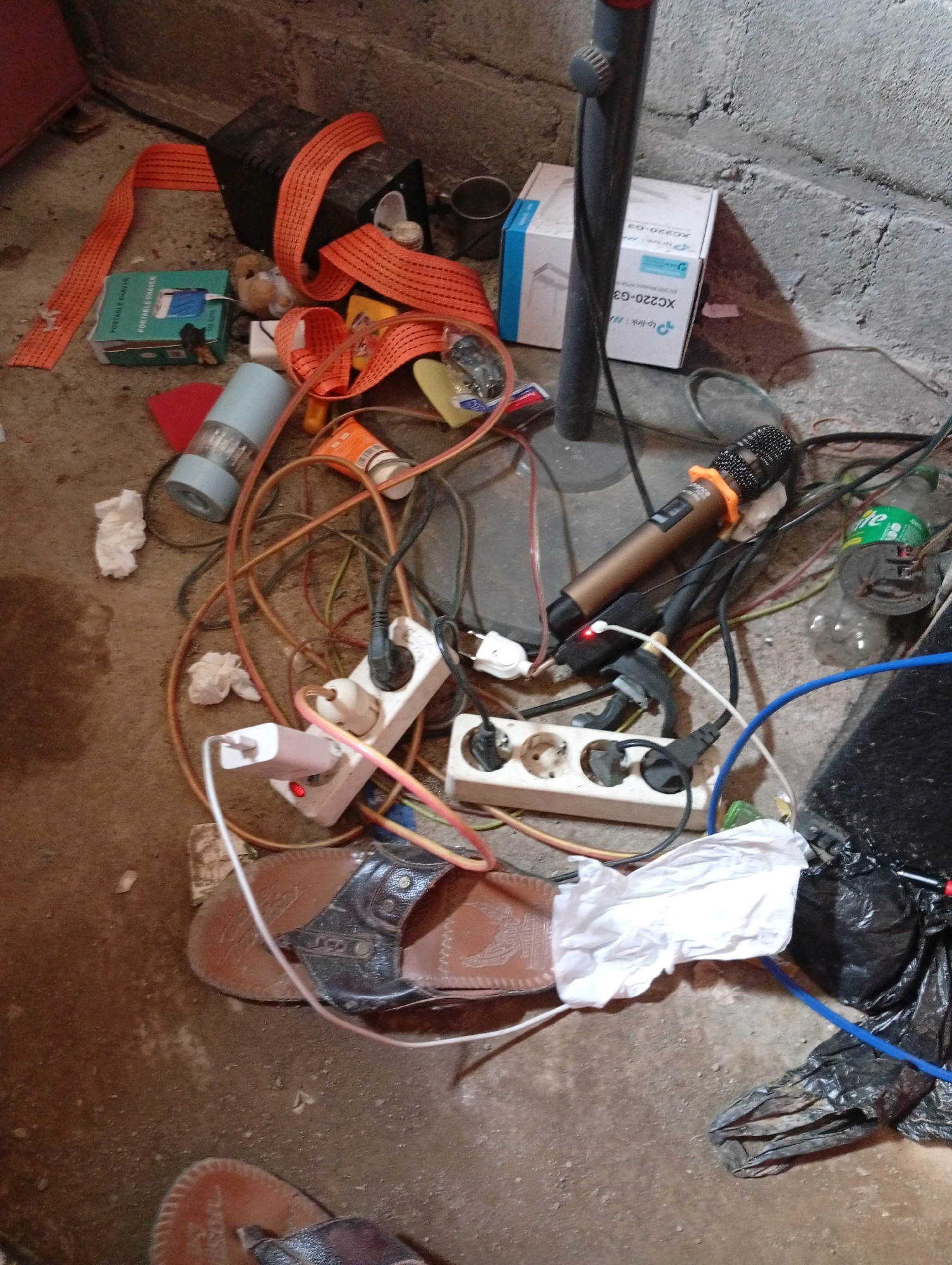 The image depicts a cluttered ground-level scene, likely in an urban environment such as Blimbing, Indonesia, characterized by a concrete floor and a rough, textured wall. Various electronic devices and everyday objects are strewn about. In the center, a white power strip with multiple outlets is connected to several electrical cords, some of which are plugged into devices. A brown microphone with an orange collar lies near the power strip. A pair of brown and black flip-flops are visible on the floor, one partially covered by a crumpled piece of white paper. Other items include a small green box, a blue and white box, a grey cylindrical object, and a black plastic bag containing what appears to be trash. An orange strap is wrapped around a dark object in the upper left corner. The overall impression is one of disarray and potential disuse.