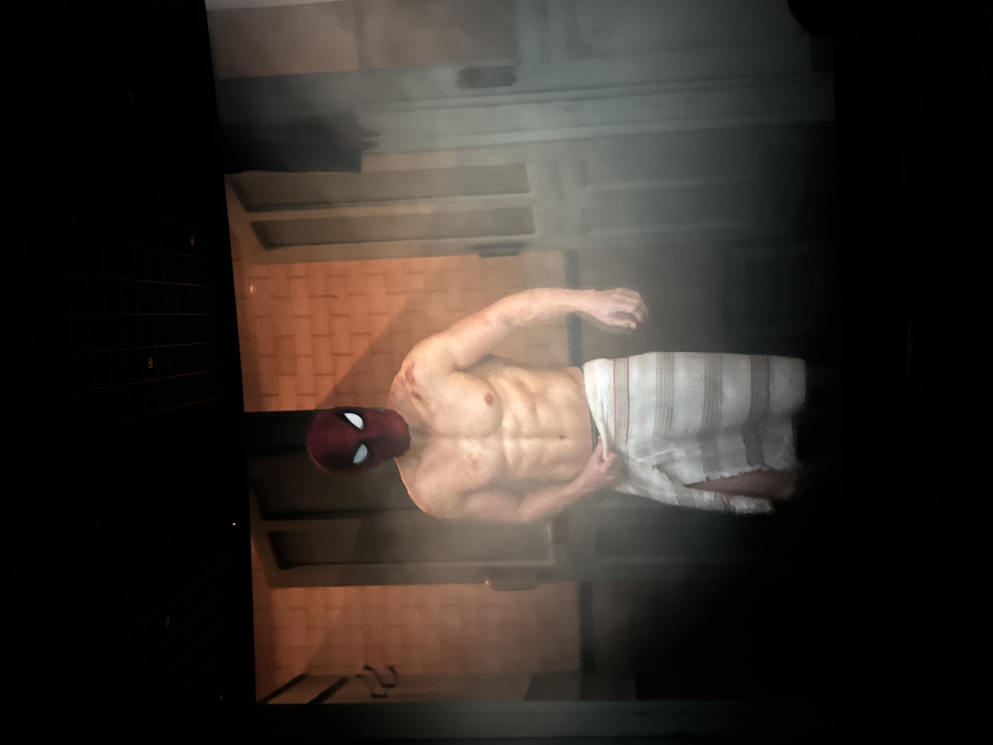 The image shows a man wearing a Spider-Man mask and a towel around his waist. He has a muscular physique and appears to be standing in a steamy or foggy environment. Behind him, there are brick walls and what look like doorways or partitions. The lighting is dim, with a warm glow emanating from behind him and the environment. The scene is dark in the foreground and edges, suggesting it might be a projection or a scene from a movie theater. There is no indication of the city of Guta, China, in this image.