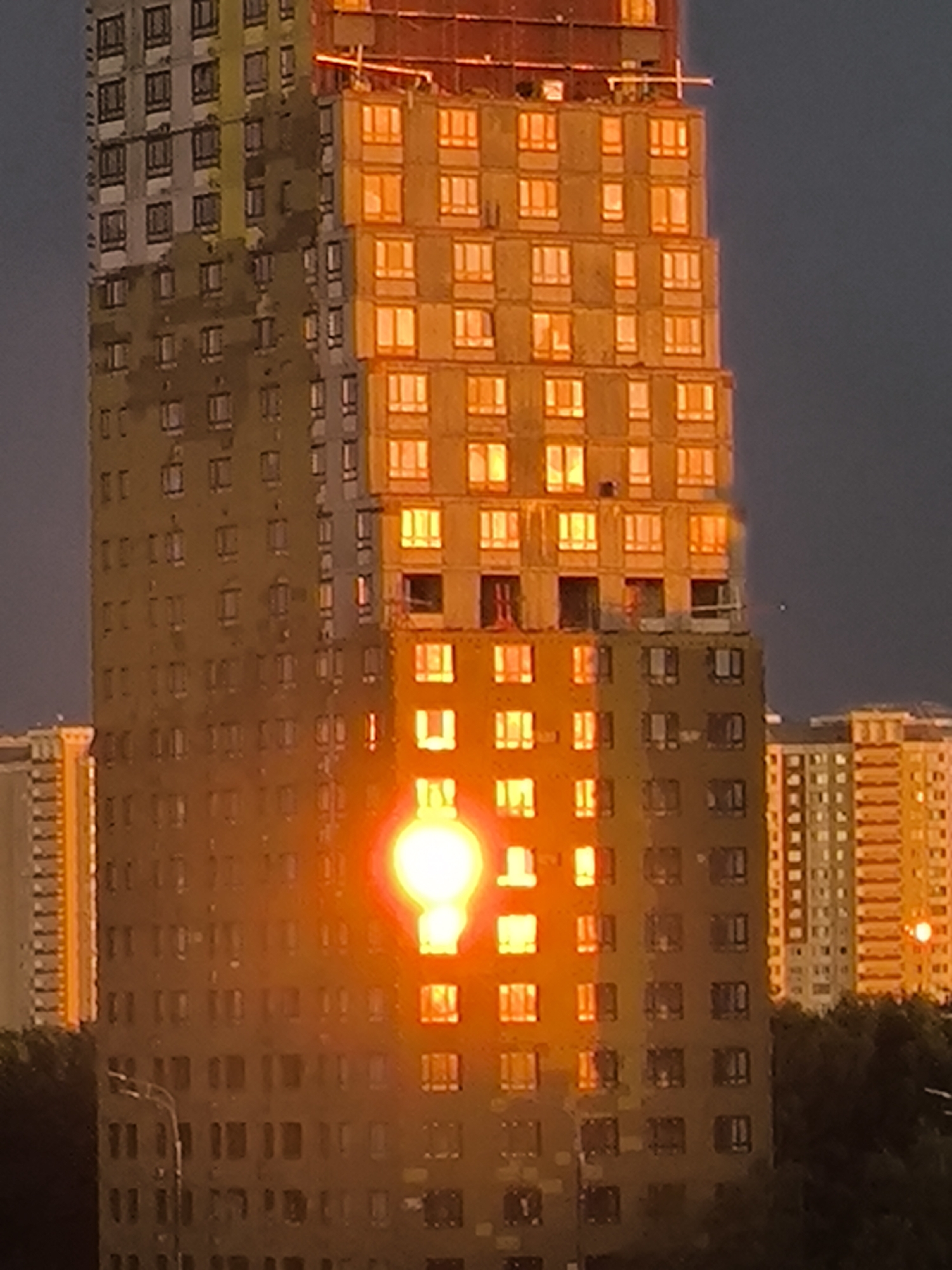 A high-angle, close-up shot captures a towering, modern skyscraper reflecting the golden hues of a setting sun against a dark, twilight sky. The building, partially under construction, exhibits two distinct sections: a left side clad in a muted, greyish-yellow material with numerous rectangular windows, and a right side adorned with a warm, orange-yellow facade, also punctuated by a grid of illuminated windows. A brilliant, circular lens flare, intensely bright and emanating a warm orange light, is positioned mid-way up the right side of the building, dominating the center of the frame. Another, smaller lens flare is visible further to the right and lower on the adjacent building. The top of the main building shows exposed structural elements, including scaffolding and rebar. In the foreground, the dark silhouettes of trees and the top of a street lamp suggest an urban environment. The overall impression is one of urban development bathed in the dramatic light of sunset.