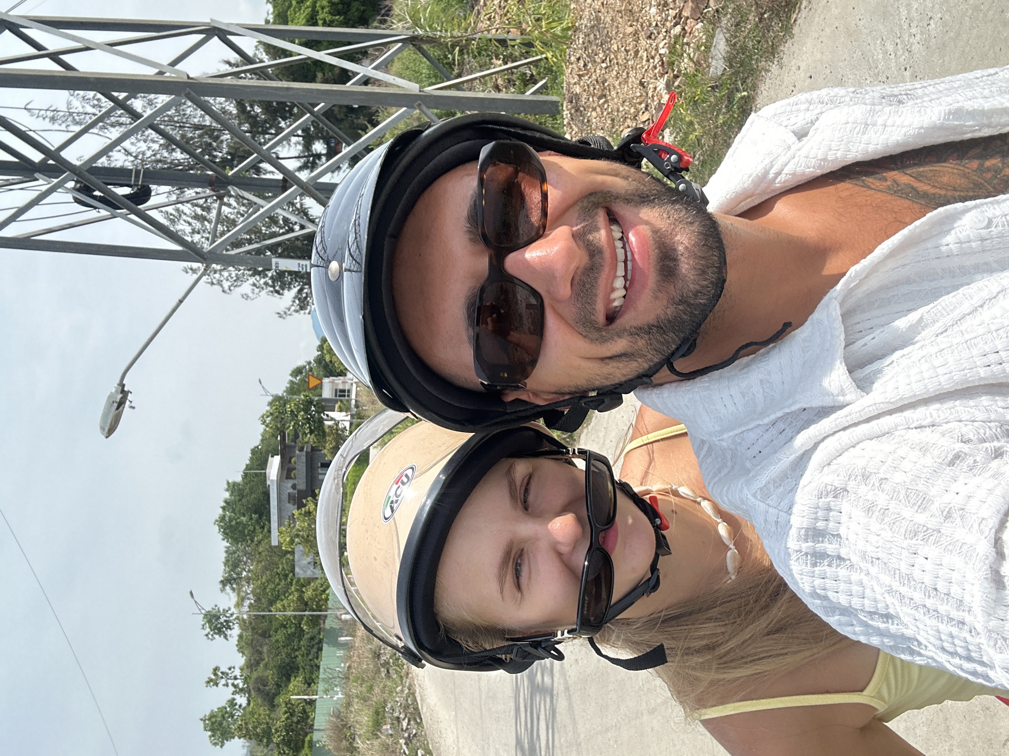 In Nam Nha Trang Ward, Vietnam, a man and a woman are taking a selfie on a motorcycle. The woman, wearing a tan helmet and a yellow tank top, has her sunglasses perched on her nose. She is also wearing a seashell necklace. The man, with a black helmet and sunglasses, is wearing a white open-collared shirt with tattoos visible on his chest. They are on a paved road, with a metal pylon and lush greenery in the background. The sky is overcast.