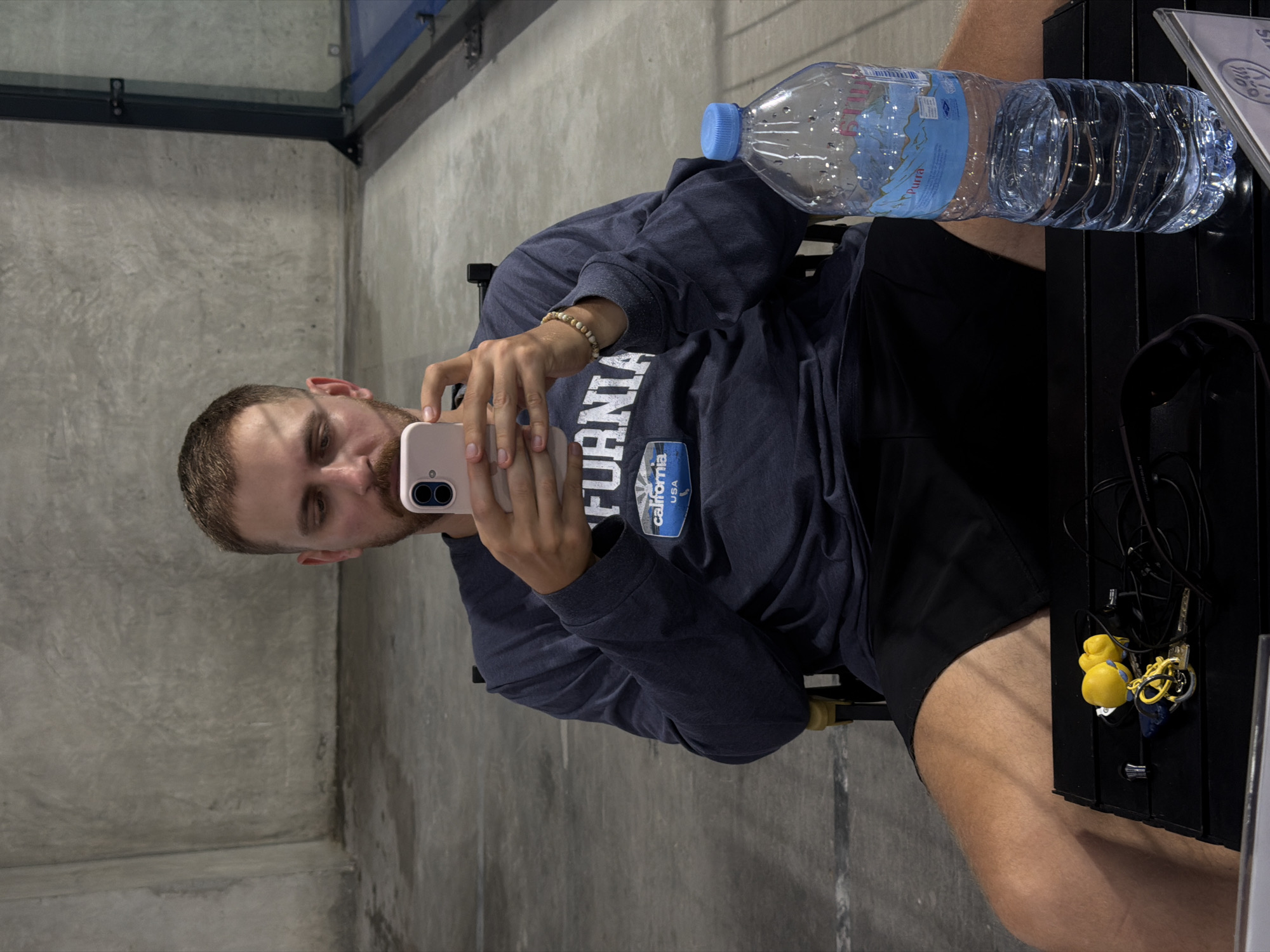 A man sits in a director's chair, looking at his pink smartphone, in front of a textured gray concrete wall. He wears a dark blue long-sleeved shirt with a "California USA" graphic and black shorts. On a black table in front of him, there's a clear plastic bottle of water with a blue cap and a label depicting mountains, along with a set of keys and earbuds. The floor appears to be concrete, and a glass partition is visible in the background on the right. The scene is likely at a sports facility, given the glass partition that could be part of a court. The provided location of Ban Buak Khrok North, Thailand, suggests this is an indoor or covered outdoor area, possibly an air-conditioned space given the man's attire.