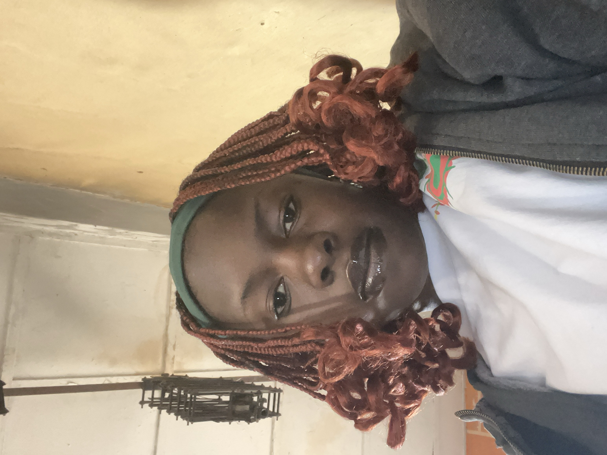 A medium shot captures a young Black woman with braided, reddish-brown hair styled with curls at the ends and a green headband. She is looking directly at the camera, her expression neutral, with dark, glossy lipstick and subtle eye makeup. She's wearing a white t-shirt with a graphic design and a grey zippered hoodie. The background is a textured, yellowish wall, and to the left, a dark metal object with a cage-like structure is visible against a light-colored, possibly ceiling, surface. The scene appears to be indoors. The overall impression is a casual, selfie-style photograph taken in a domestic or informal setting.