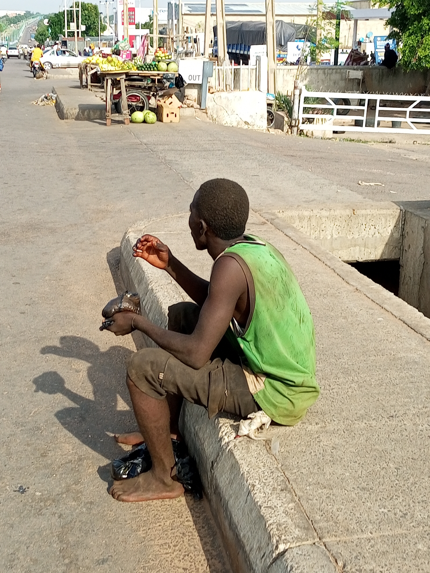 The image depicts a young Black male sitting on a concrete curb at the side of a road in Tempure, Nigeria. He is wearing a stained green sleeveless shirt and brown shorts, with his bare feet resting on a black plastic bag. In his right hand, he is holding some small items, possibly food, up to his mouth. He appears to be looking off to the side, away from the camera.
The surrounding environment includes a paved road with tire marks, suggesting it is a traffic area. In the background, a fruit stall is visible with various produce like bananas, watermelons, and cucumbers displayed on wooden carts. Further in the background, there are buildings, some appearing to be shops or stalls, and a few vehicles, including a yellow motorcycle and a white car. The overall scene suggests a bustling urban environment, likely a market area on the roadside. A large concrete structure with an opening is visible to the right of the seated individual.
