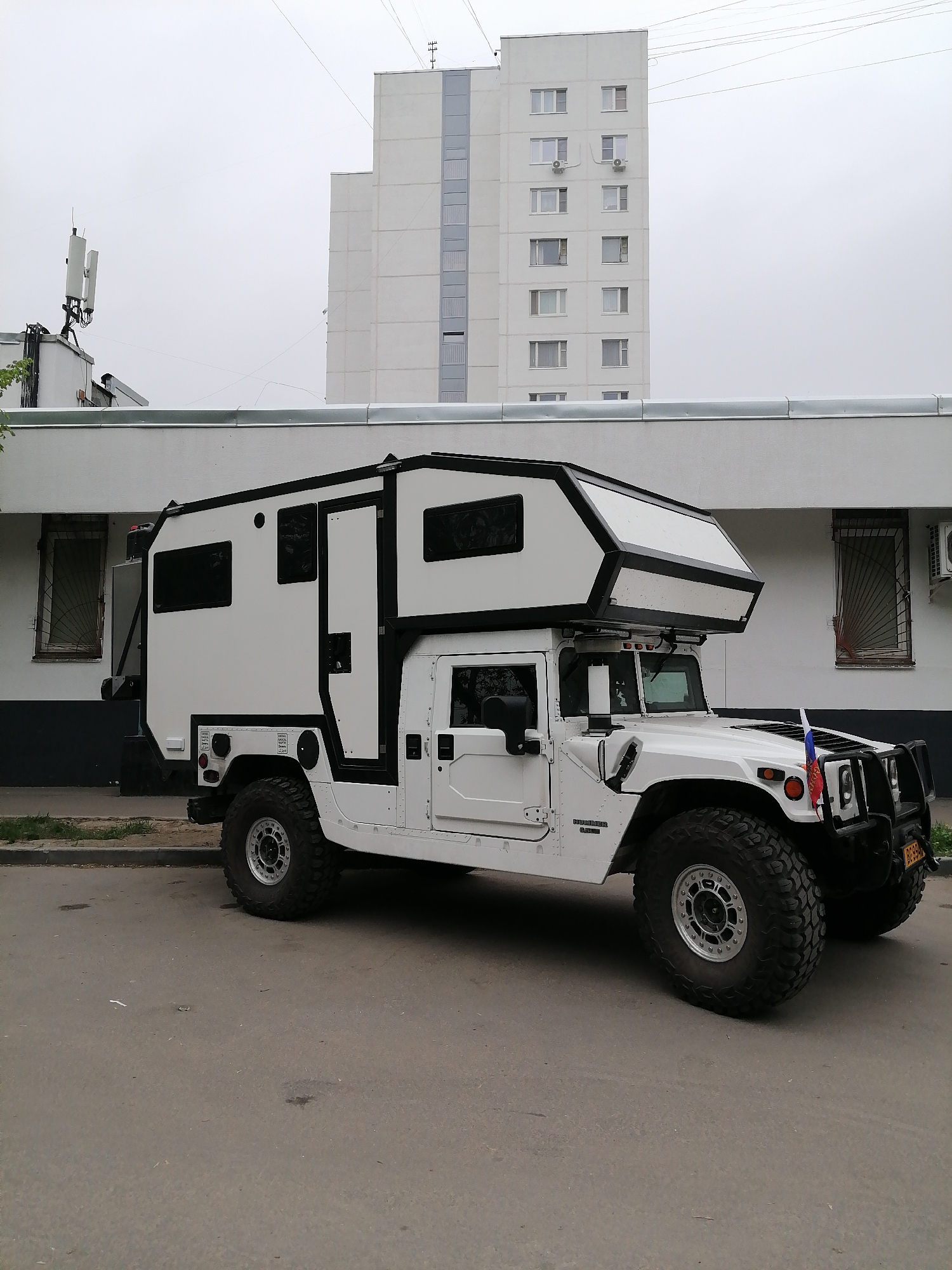 A white Hummer H1 vehicle, modified with a camper shell, is parked on a paved road next to a low, white building. The vehicle has a Russian flag attached to the front bumper. A taller, white apartment building is visible in the background. The location is Moscow, Russia. The sky is overcast.