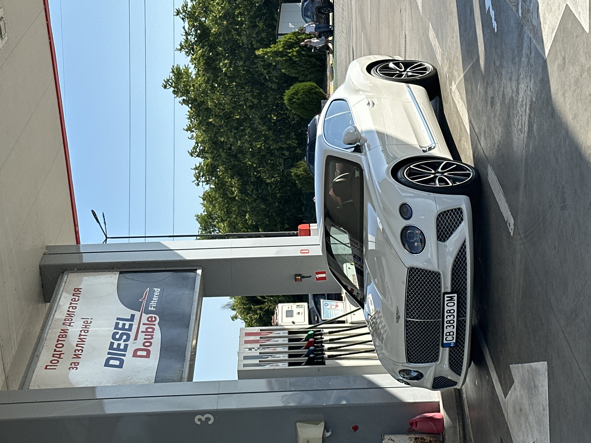 A white Bentley Continental GT is parked at a gas station. The car's license plate reads "CB 3838 OM". In the background, there is a sign with text in Bulgarian, "Подготви двигателя за излитане!" which translates to "Prepare the engine for takeoff!". The gas station appears to be located in Polsko Padarevo, Bulgaria, as suggested by the surrounding landscape of trees and the overall feel of a Bulgarian gas station. People are visible in the background, walking near other vehicles. The sky is clear and blue, indicating a sunny day. The ground is asphalt, with white painted lines marking parking spaces and directional arrows.