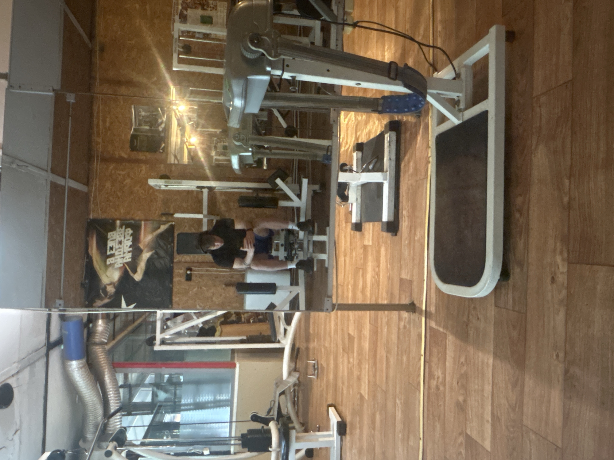 The image depicts a gym interior, with various exercise machines present. A man is visible, seated on a machine, possibly engaged in a workout. Reflective surfaces create a mirrored effect, doubling the visual elements. The surroundings include walls with posters and ventilation ducts. The gym's flooring has a wood-like appearance. While the image is from a gym, there is no indication of its location within Elhovo, Bulgaria.