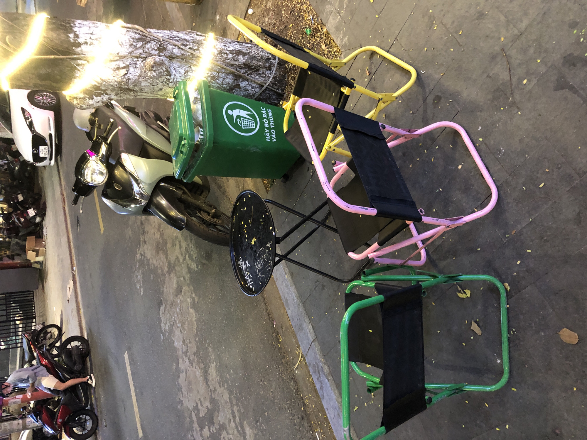 The image depicts an outdoor scene in Da Nang, Vietnam, likely a sidewalk or street area. A red scooter with a person on it, wearing a blue shirt and white shorts, is parked on the left. The person is looking at their phone. To the right of the scooter, a silver scooter is parked, and a green trash bin with Vietnamese text and a recycling symbol is visible. A tree adorned with string lights is on the right side of the frame. In the foreground, three folding chairs are arranged around a black, foldable table. Two of the chairs are colorful: one is bright yellow, and the other is pink. A third chair, in the foreground left, has a green frame. The ground is paved, and scattered leaves are visible. In the background, a white car and other parked motorcycles are present, along with a building structure. The overall impression is of a casual outdoor seating area, possibly associated with a nearby establishment.