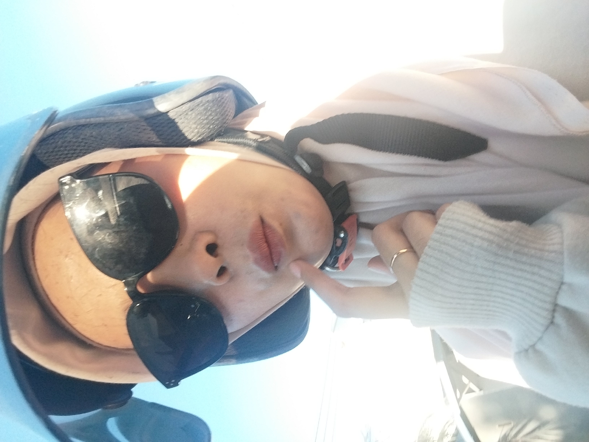 A selfie, taken by a person wearing a blue helmet and black sunglasses, shows them pointing a finger to their chin. The person's mouth is slightly pursed, and they are wearing a light-colored hijab and a white sweater. A black strap from the helmet is visible around the neck. The background is a bright, blown-out sky, suggesting an outdoor setting in a sunny location. The location is identified as Cinnong, Indonesia.