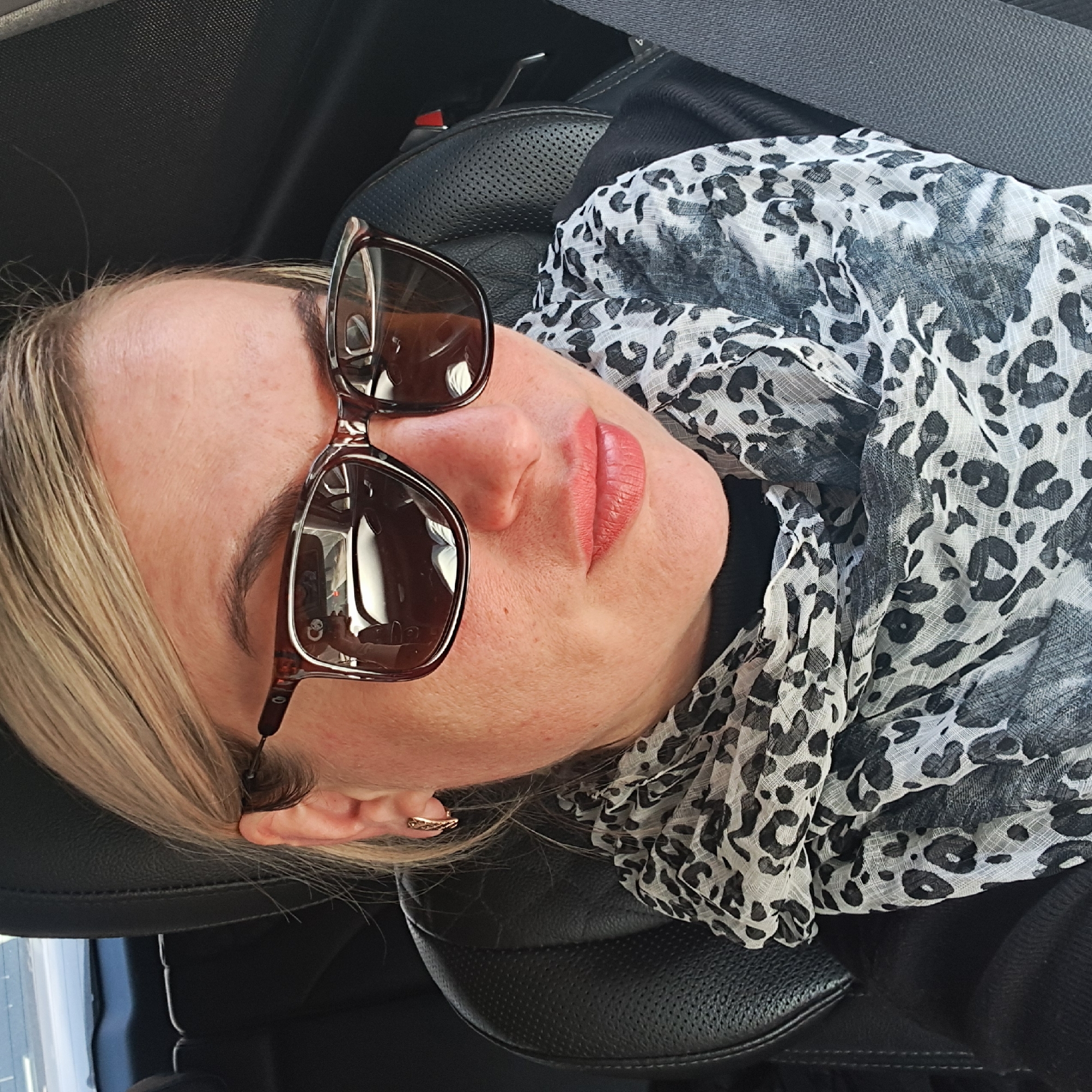 A middle-aged, fair-skinned woman is reclining in a black leather car seat. She is wearing brown-framed sunglasses perched on her nose, and a black and white leopard-print scarf is draped around her neck and chest. She has blonde hair and a small gold hoop earring in her left ear. The interior of the car is visible in the background, including a seatbelt. The location in Elektrougli, Russia, cannot be confirmed from the image.