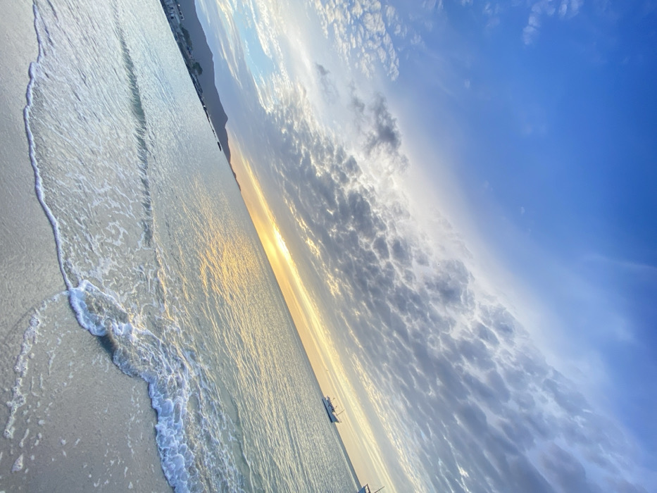 The image displays a coastal landscape in Juan Griego, Venezuela, captured during either sunrise or sunset. The foreground features a light-colored sandy beach with white foam lines from gentle, receding waves. The midground shows calm, light-blue ocean water reflecting a golden glow from the sun, which is low on the horizon. Two boats are present on the water's surface; one appears as a prominent white fishing vessel positioned to the right of the sun's reflection, and a second, smaller vessel is faintly visible further right on the horizon. To the distant left, a landmass characterized by dark mountains or hills is visible, with subtle indications of structures along the shoreline. The sky occupies the upper portion of the frame, transitioning from a clear, lighter blue with scattered wispy clouds in the upper left to a denser formation of grey and white altocumulus-like clouds, illuminated from below by the sun's light, stretching across the middle and lower right sky. No people are visible in the scene.
