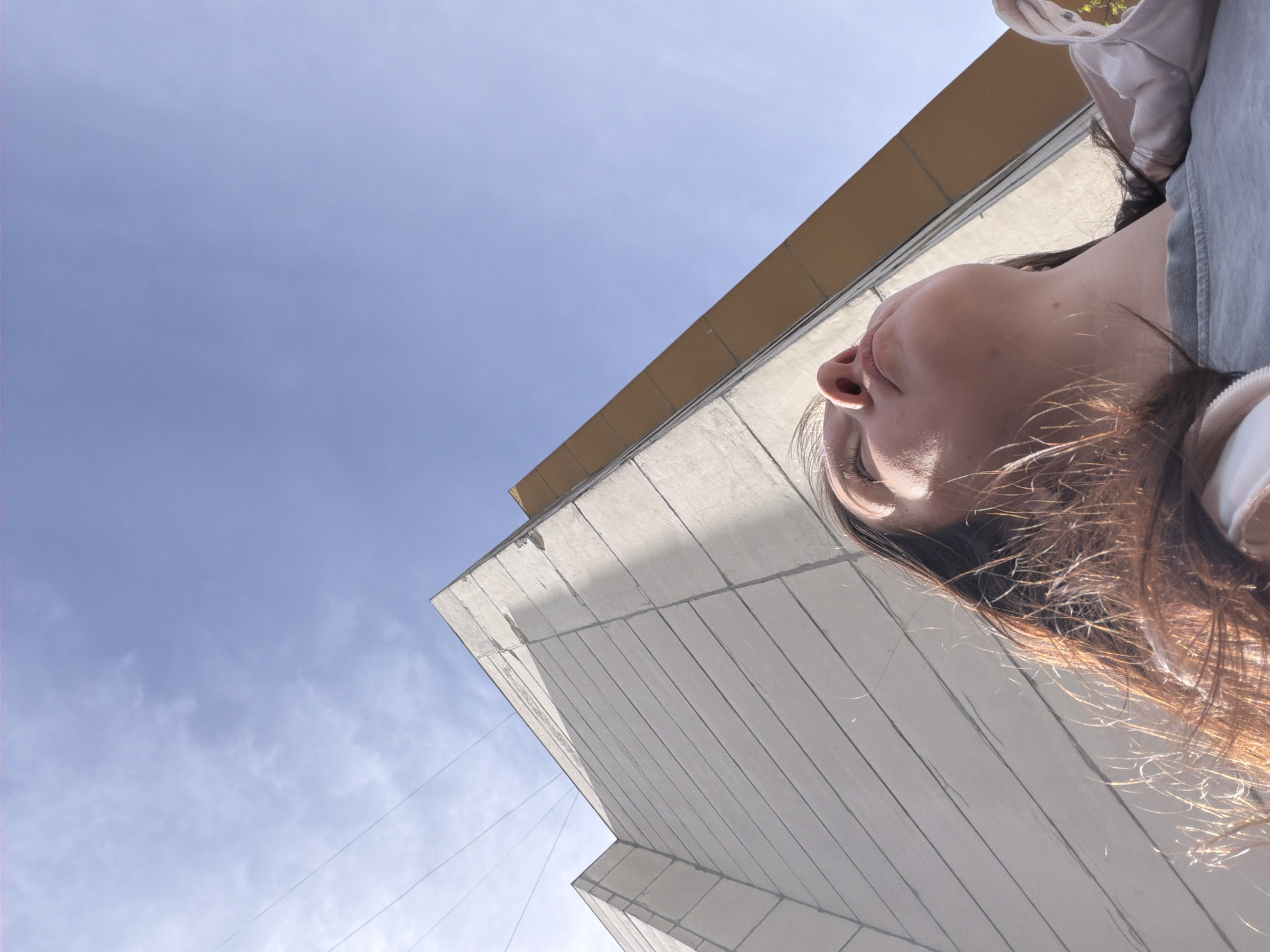 A low-angle shot, likely taken from below, depicts a woman looking upwards at a building and the sky. The sky is a clear, pale blue with faint wisps of clouds visible in the upper left portion of the frame. The building is a modern-looking structure, possibly made of concrete, with a horizontal gold-colored overhang. The woman's face is partially visible, with her chin, nose, and one eye prominent. She has brown hair that is catching the sunlight. She is wearing a light-colored jacket or shirt. The scene takes place outdoors, likely in an urban setting. There is no evidence to suggest that this image was taken in Staraya, Russia.