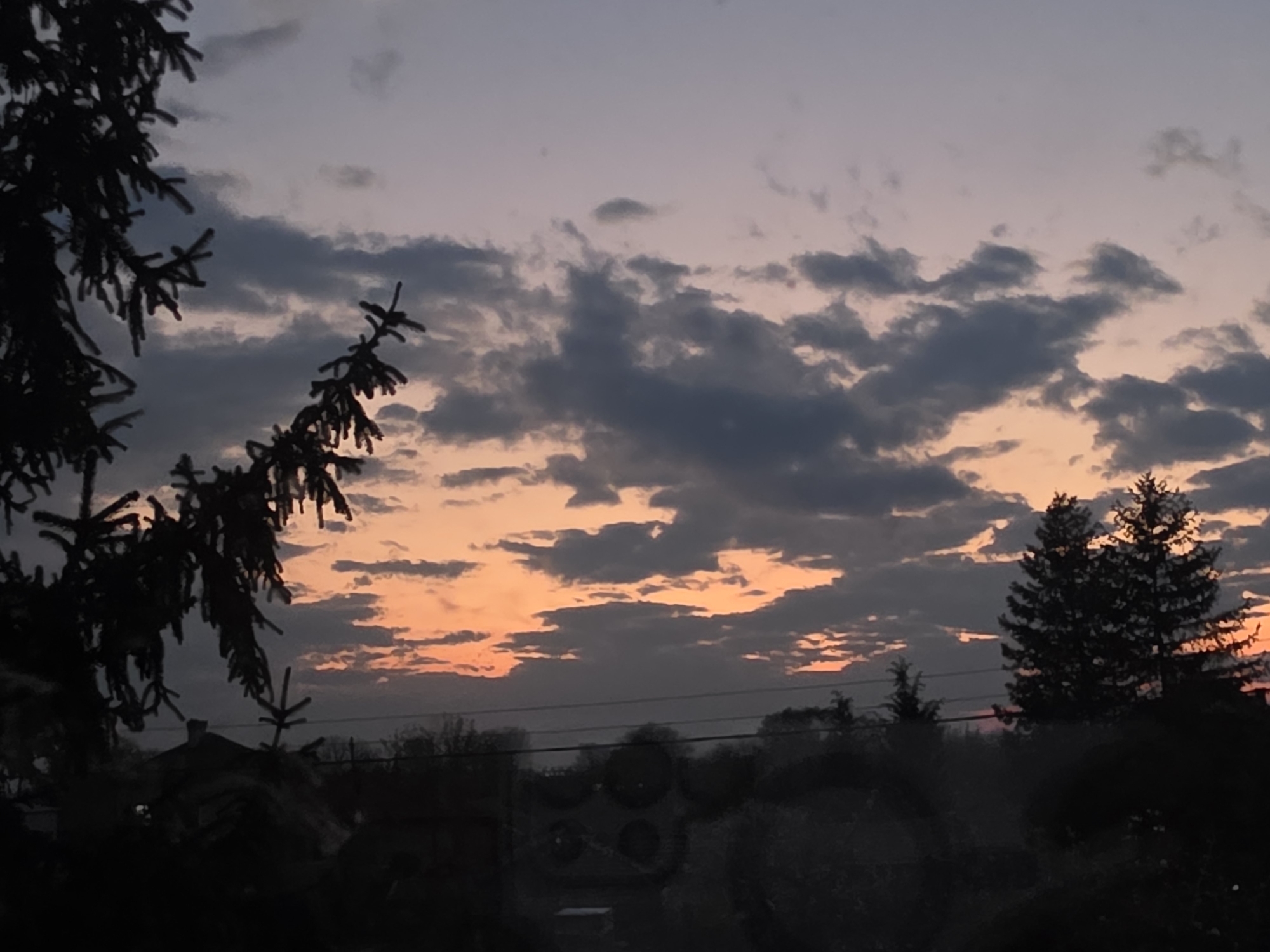 The image captures a twilight sky over Yagodovo, Bulgaria. The sky transitions from a light purple hue at the top to orange near the horizon, with scattered grey clouds. Silhouetted trees frame the image on both sides. The background shows a blurry, dark outline of buildings and trees. No people or interactions are visible in the image.