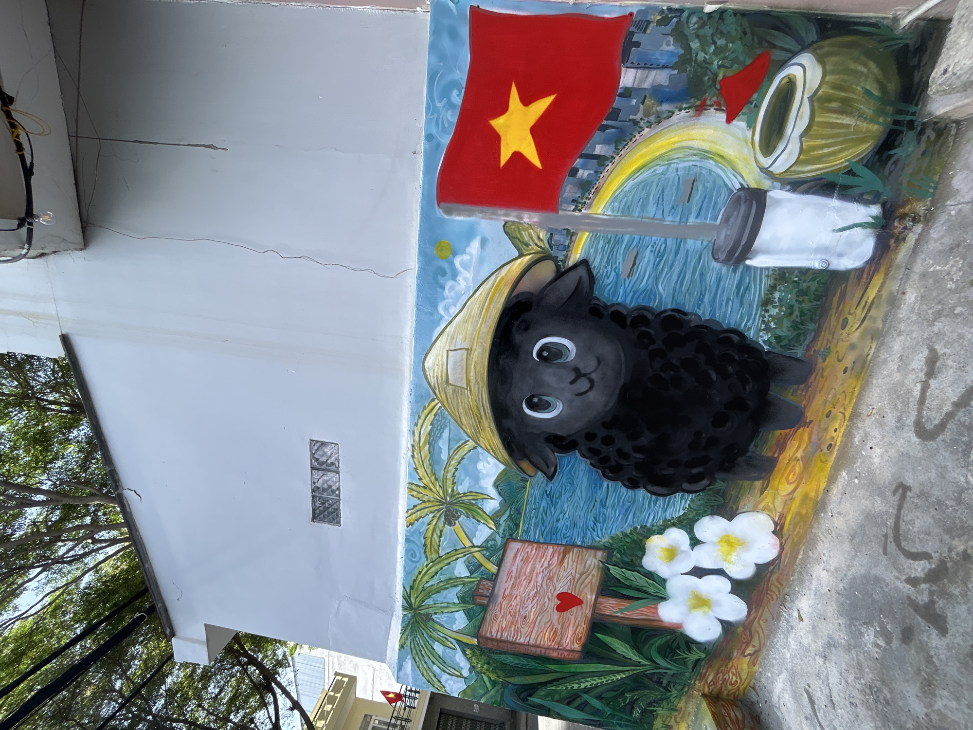 A brightly painted mural covers a section of an outdoor wall in Bac Nha Trang Ward, Vietnam. The mural features a black sheep wearing a conical hat, positioned in front of the Vietnamese flag. Behind the flag, the cityscape of Nha Trang is faintly visible. Other elements of the mural include palm trees, a coconut, a cup, wooden signage with a heart on it, and white flowers. The mural is on a concrete surface next to a building facade.