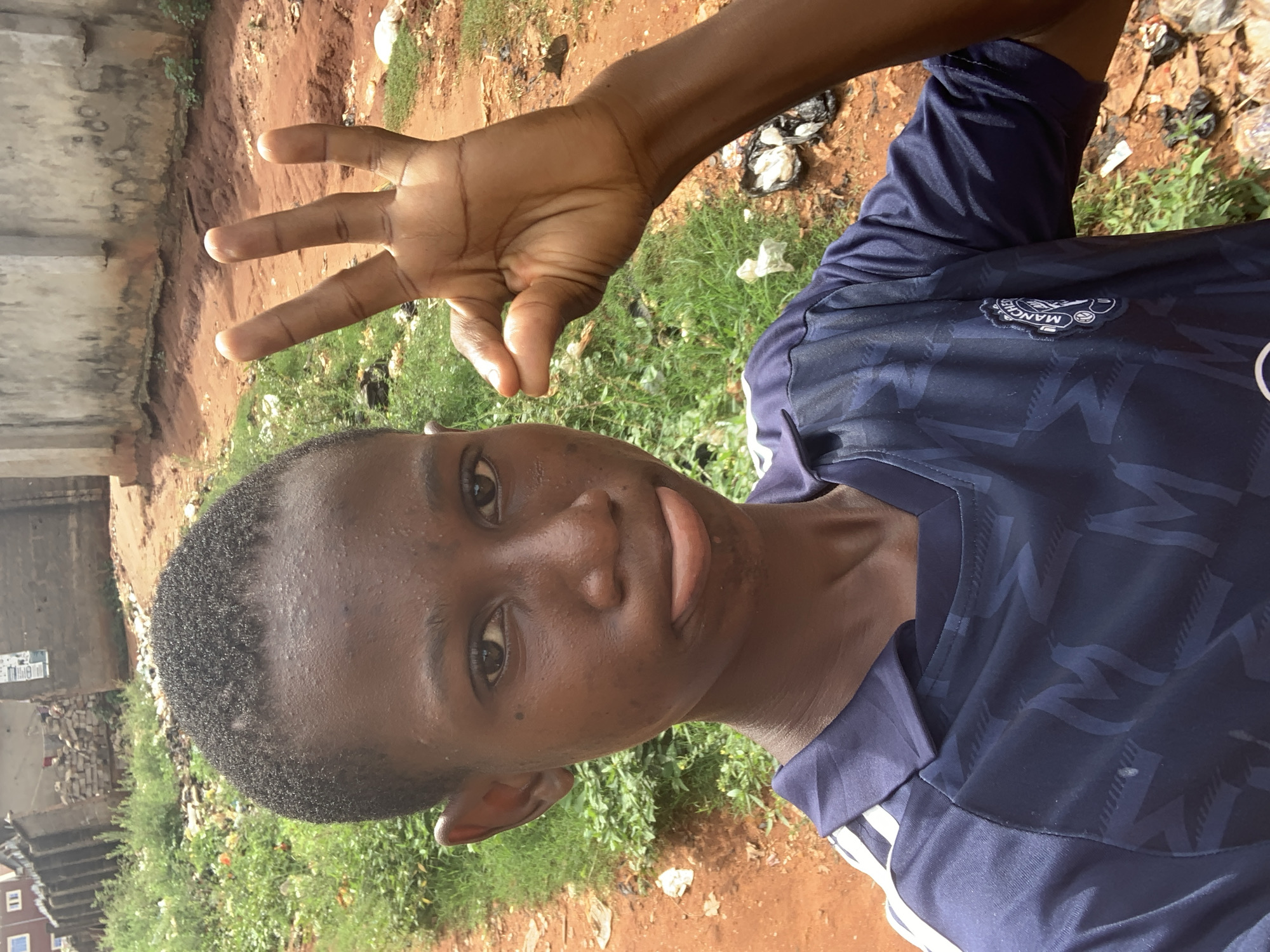A young person with short, dark hair is holding up three fingers. They are wearing a blue and navy Manchester United soccer jersey with white stripes. The background appears to be an outdoor area in Onitsha, Nigeria, with a mix of dirt, grass, and litter. In the distance, there is a cement structure and some brick buildings.