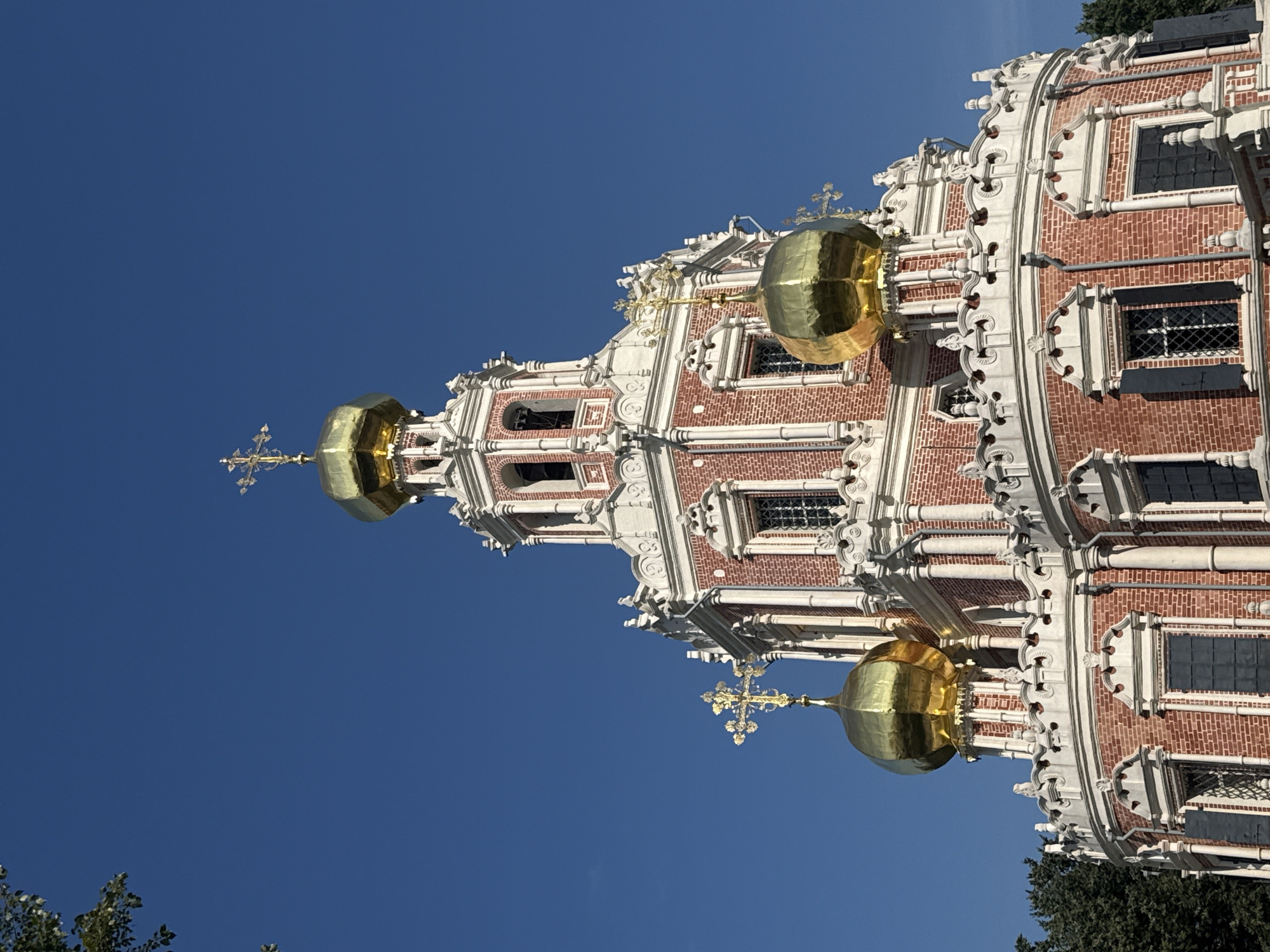The image shows the Church of the Intercession at Fili, located in Moscow, Russia. The church, a prominent example of Moscow Baroque architecture, is predominantly red brick with white decorative elements. It features a central, multi-tiered structure topped with a golden dome and cross. Smaller golden domes with crosses are visible on lower sections of the church. The building is set against a clear, bright blue sky, and on the left side of the frame, the leafy branches of a tree are partially visible. The lower portion of the church is circular with multiple windows.