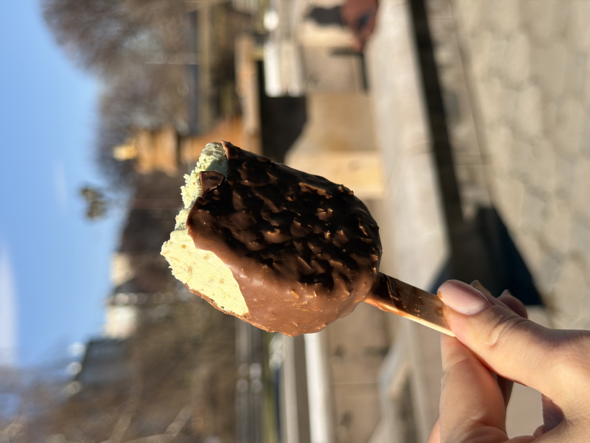 A close-up, portrait-oriented image shows a hand holding a partially eaten ice cream bar on a bright, sunny day in Krasnoyarsk, Russia. The ice cream itself has a light green or pistachio-colored interior, which is visible where a bite has been taken from its upper left side. It is covered in a thick, dark brown chocolate shell that appears to contain small pieces, likely nuts or crisps, giving it a textured, bumpy finish. The ice cream is attached to a simple wooden stick, which bears a very faint, unidentifiable circular mark or logo near where it is held.
The hand holding the ice cream has short, neatly manicured nails painted in a subtle light pink or nude color. Warm, natural sunlight illuminates the ice cream and hand, casting soft highlights and shadows.
The background is significantly blurred, creating a shallow depth of field that emphasizes the ice cream. Visible elements in the soft-focus background include a clear blue sky on the left, indicating good weather. To the right and below, there are hints of blurred trees and architectural structures, suggesting an outdoor, urban setting such as a park or city street. The bright lighting conditions indicate it is daytime, contributing to a pleasant and leisurely atmosphere. No legible text is visible in the image.