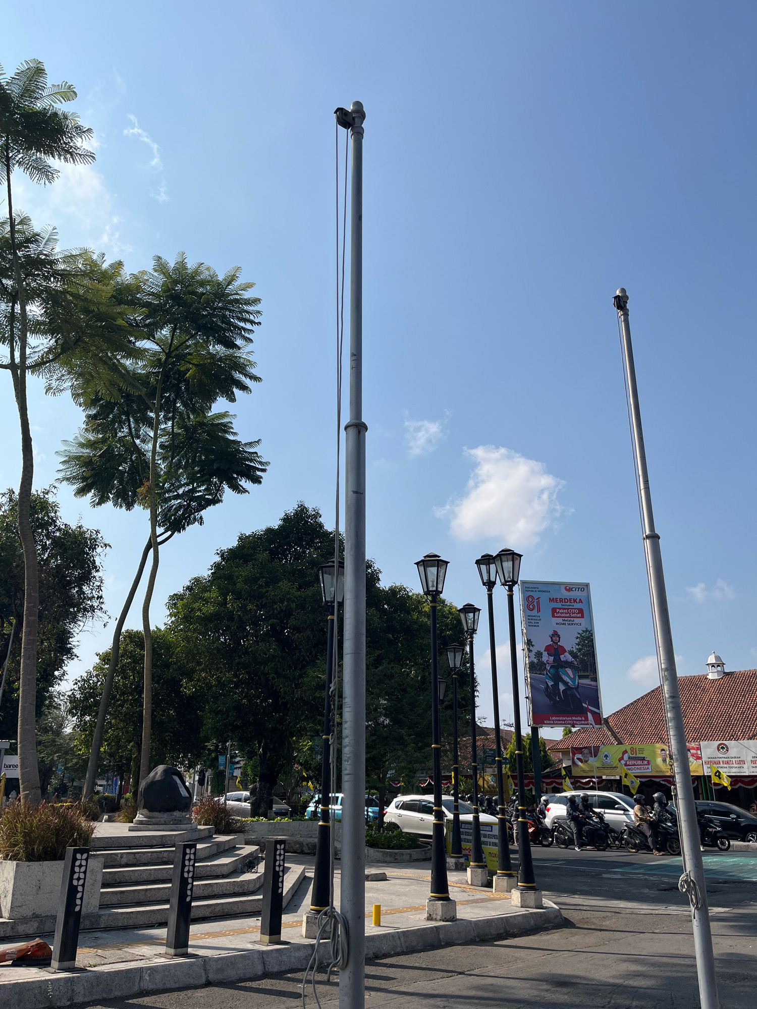 The image depicts a street scene in Yogyakarta, Indonesia, under a clear blue sky with scattered clouds. In the foreground, a paved walkway with bollards separates the street from a slightly elevated plaza area. The plaza features a tiered stone structure with a large dark rock monument at its center, surrounded by planters with dry grass. Several streetlights with ornate black bases and glass lanterns line the walkway. Tall, slender palm-like trees and dense green foliage create a canopy overhead. A tall metal pole, likely a utility pole or flagpole, dominates the right side of the frame, with a camera or speaker mounted at its top.
In the middle ground, a busy street is visible with a line of motorcycles and several cars, including white sedans and a darker vehicle. A large billboard advertising "CITO MERDEKA" featuring a person on a scooter is prominently displayed. Beyond the street, buildings with reddish-brown tiled roofs and white facades are visible, suggesting commercial or institutional structures. The overall impression is that of a public urban space with elements of greenery, street furniture, and commercial activity.