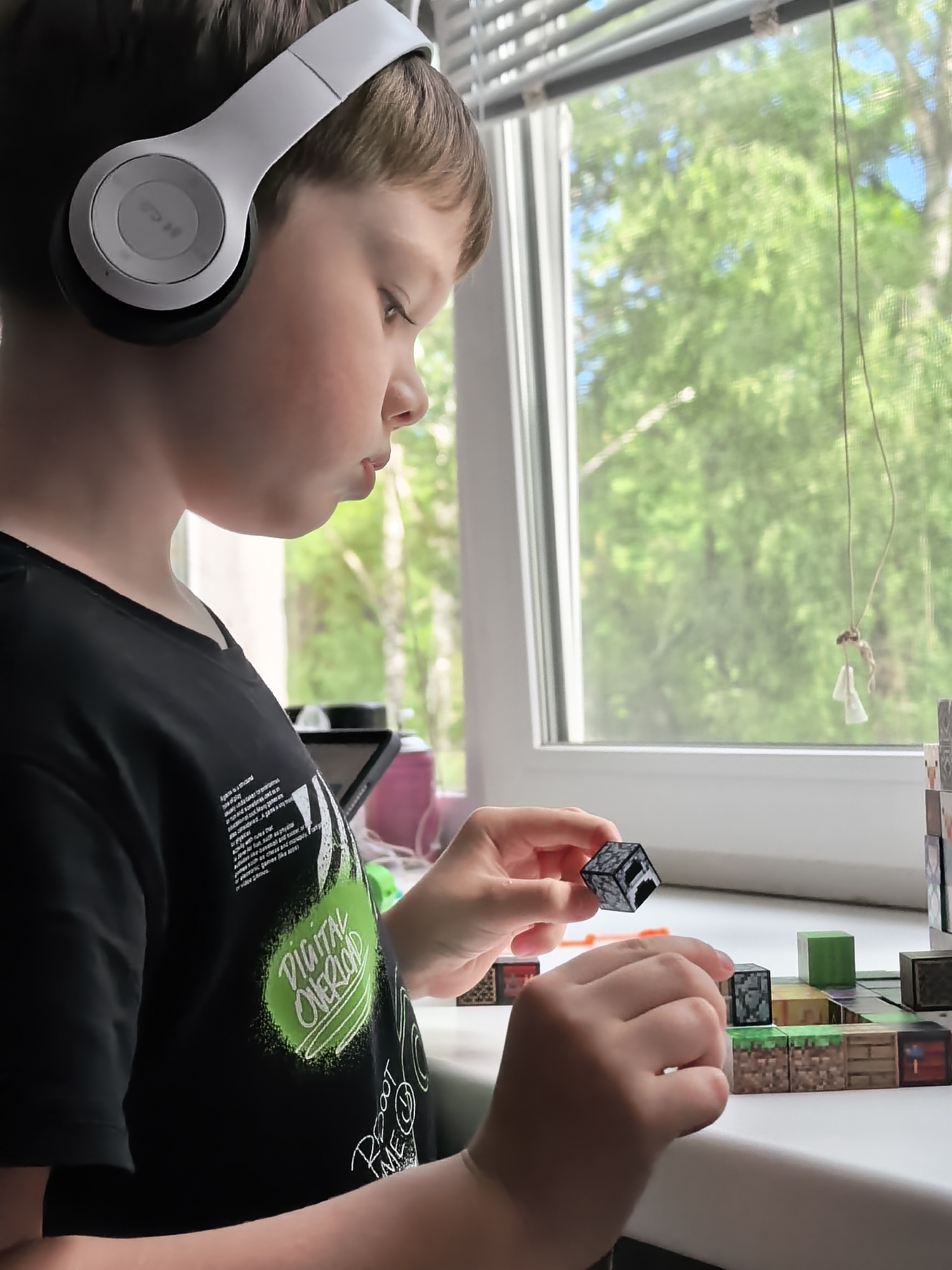 A young boy wearing white headphones is sitting at a windowsill, looking down at a game board made of Minecraft-themed blocks. He is holding a small block in his right hand, and his left hand is placed on the game board. The game board appears to be set up on the windowsill, with various blocks representing different terrains and structures. The boy is wearing a black t-shirt with a green graphic on it that reads "DIGITAL OVERLOAD". In the background, a window looks out onto green trees and a blue sky. The lighting suggests it is daytime.