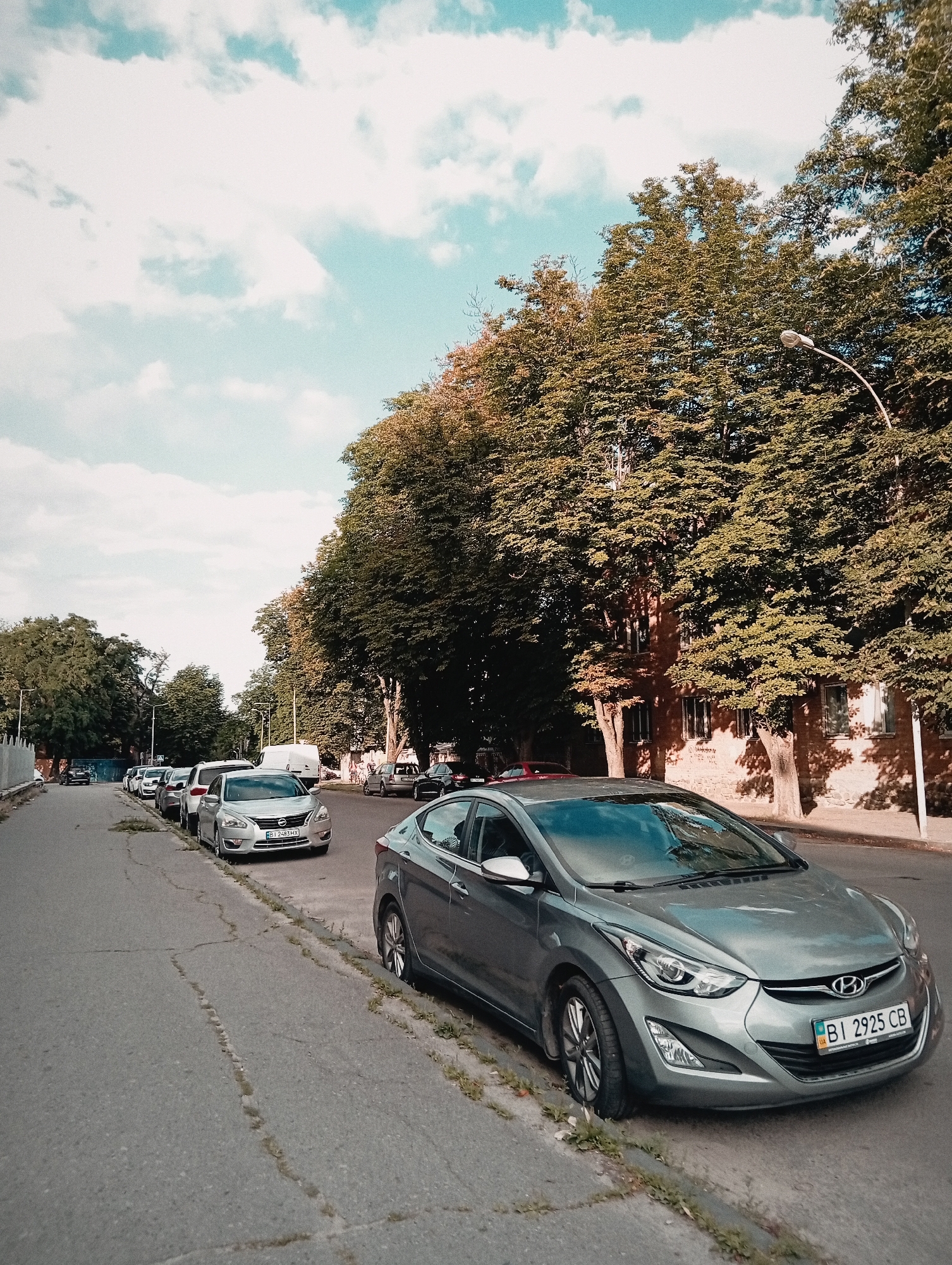 A medium shot captures a street scene in an urban environment, possibly a residential area, with a row of parked cars on the left side of the road and a building with trees in front of it on the right. The foreground is dominated by the front portion of a silver Hyundai Elantra, parked parallel to the curb. The car's license plate is visible and reads "BI 2925 CB". Several other cars are parked further down the street to the left, receding into the distance. The road surface appears aged, with visible cracks. Mature trees with dense foliage line the right side of the street, casting shadows on the sidewalk and the building behind them. The building is a multi-story structure with visible windows and a brick facade. The sky above is partly cloudy with patches of blue. A street lamp is visible in the upper right portion of the frame.