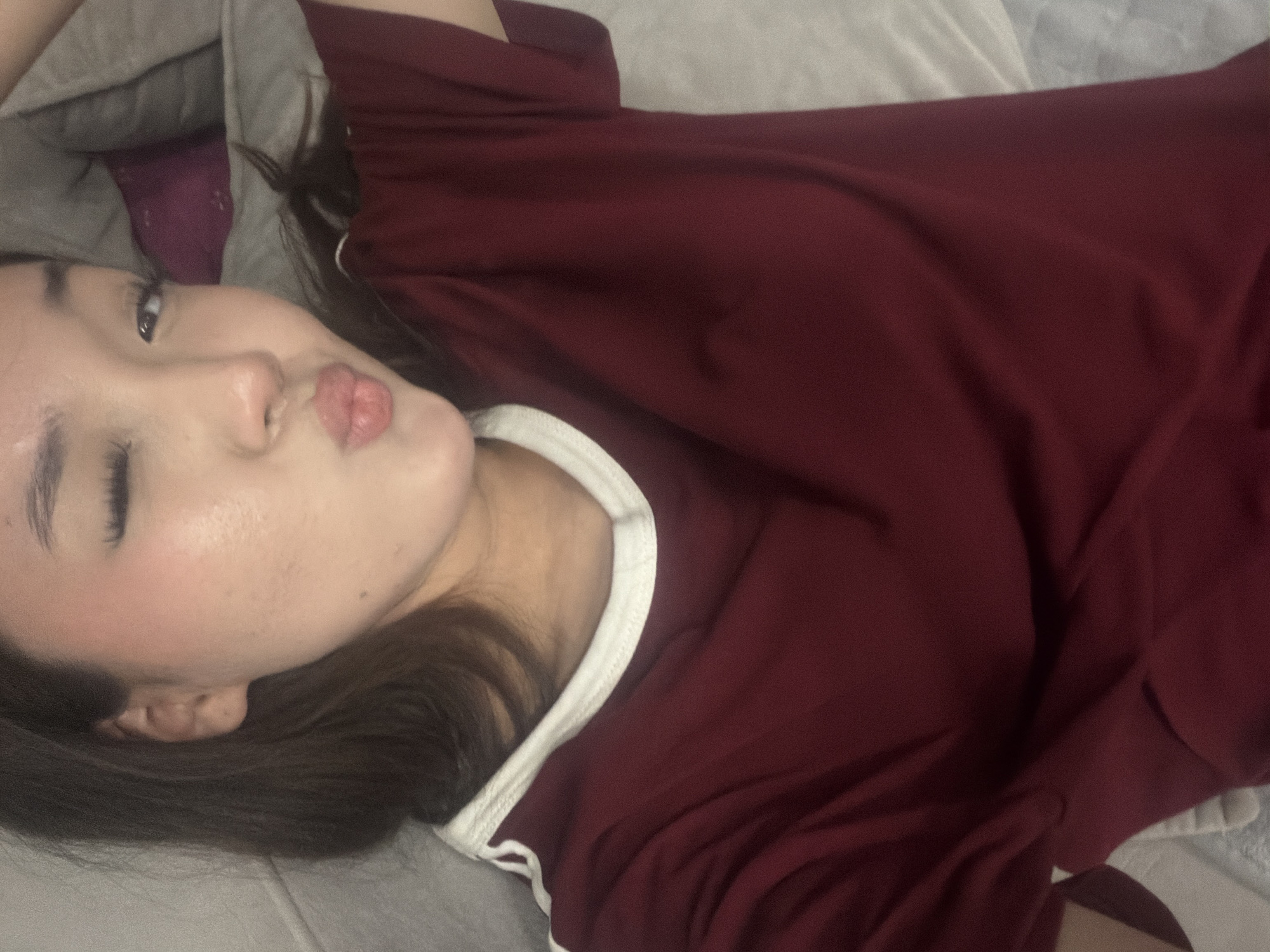 An indoor, medium shot shows a young East Asian woman with dark hair, wearing a dark red t-shirt with a white trim, lying down on a bed. She is winking with her right eye and puckering her lips. The background consists of pillows and blankets, suggesting a bedroom environment. No city or location information is discernible from the image.