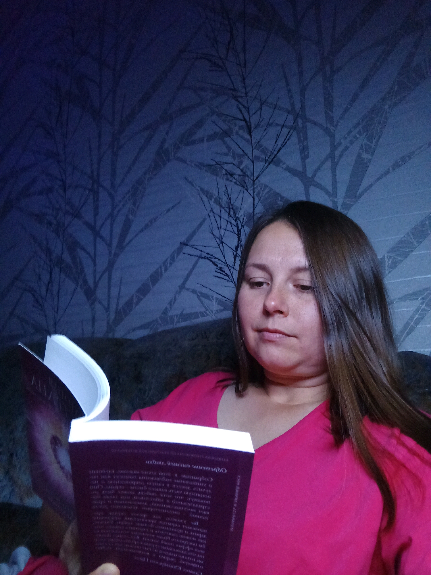 A medium shot captures a woman with long brown hair, wearing a bright pink v-neck shirt, engrossed in reading a book. She is seated, with her head tilted downwards towards the book, her expression suggesting concentration. The book, held open in her hands, has a dark purple cover with white text. The background features a dark blue wallpaper with a subtle pattern of bare tree branches. The lighting is dim, creating a moody atmosphere. The image appears to be taken indoors, likely in a residential setting. There are no visible clues to pinpoint the specific location within Novosibirsk, Russia.