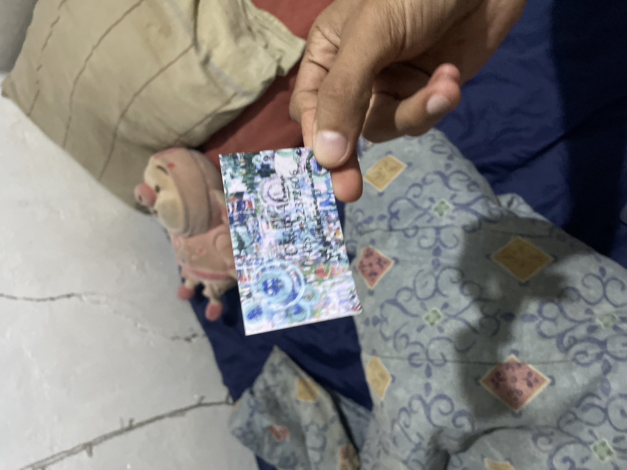 A hand holds up a small, rectangular card with a colorful, abstract design. The card appears to be a gift card or promotional item. In the background, a stuffed animal, possibly a pig, lies on a bed with blue and gray patterned sheets and a beige pillow. The lighting suggests an indoor setting, possibly a bedroom. The scene is unfocused and appears to be taken with a mobile phone.
