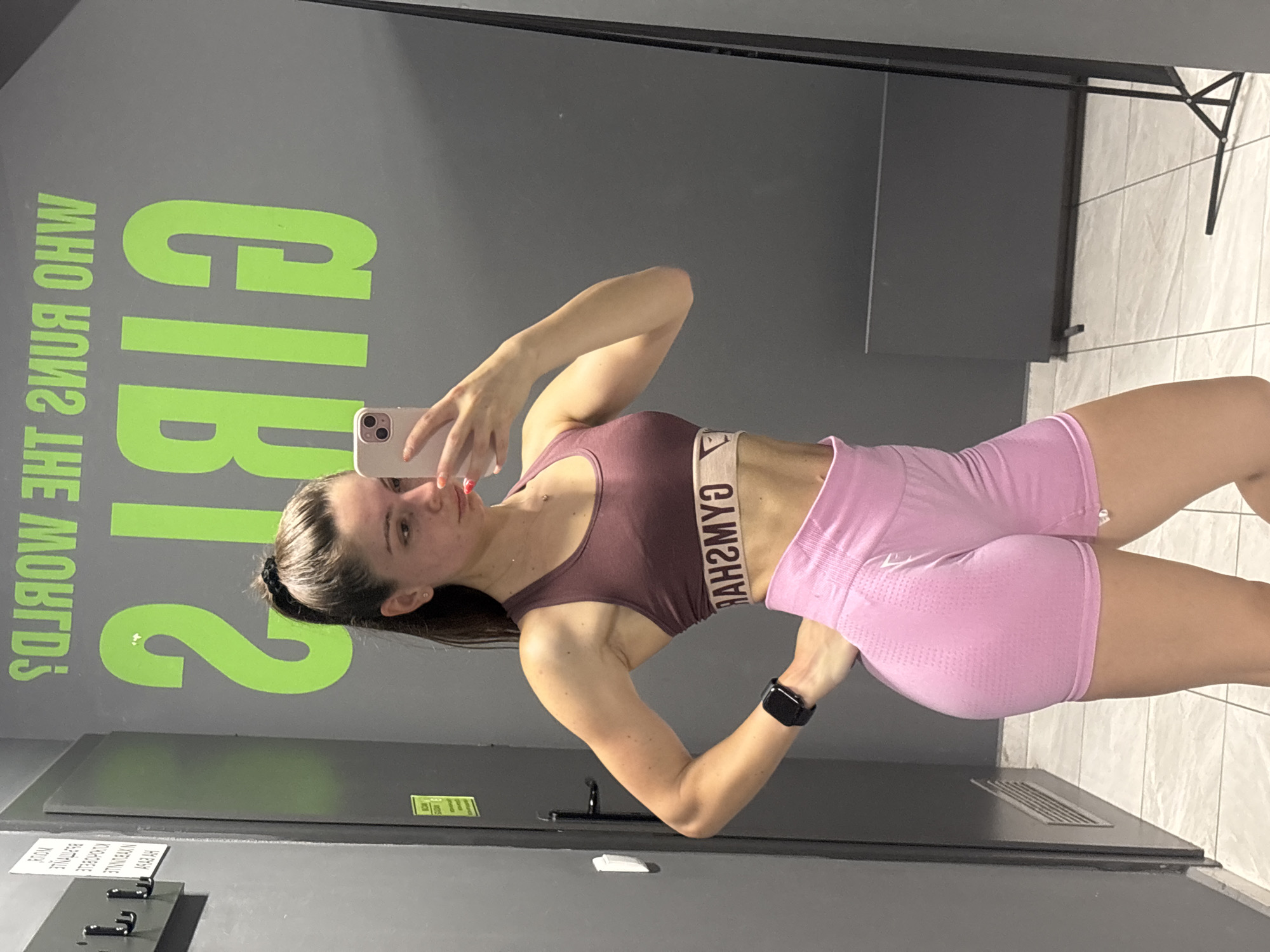 A woman is taking a selfie in a gym. She is wearing a maroon sports bra and pink shorts. The gym has gray walls with green lettering that says "GIB". There is a metal coat rack on the wall. The location is likely in the city of Varna, Bulgaria, based on the prompt request.