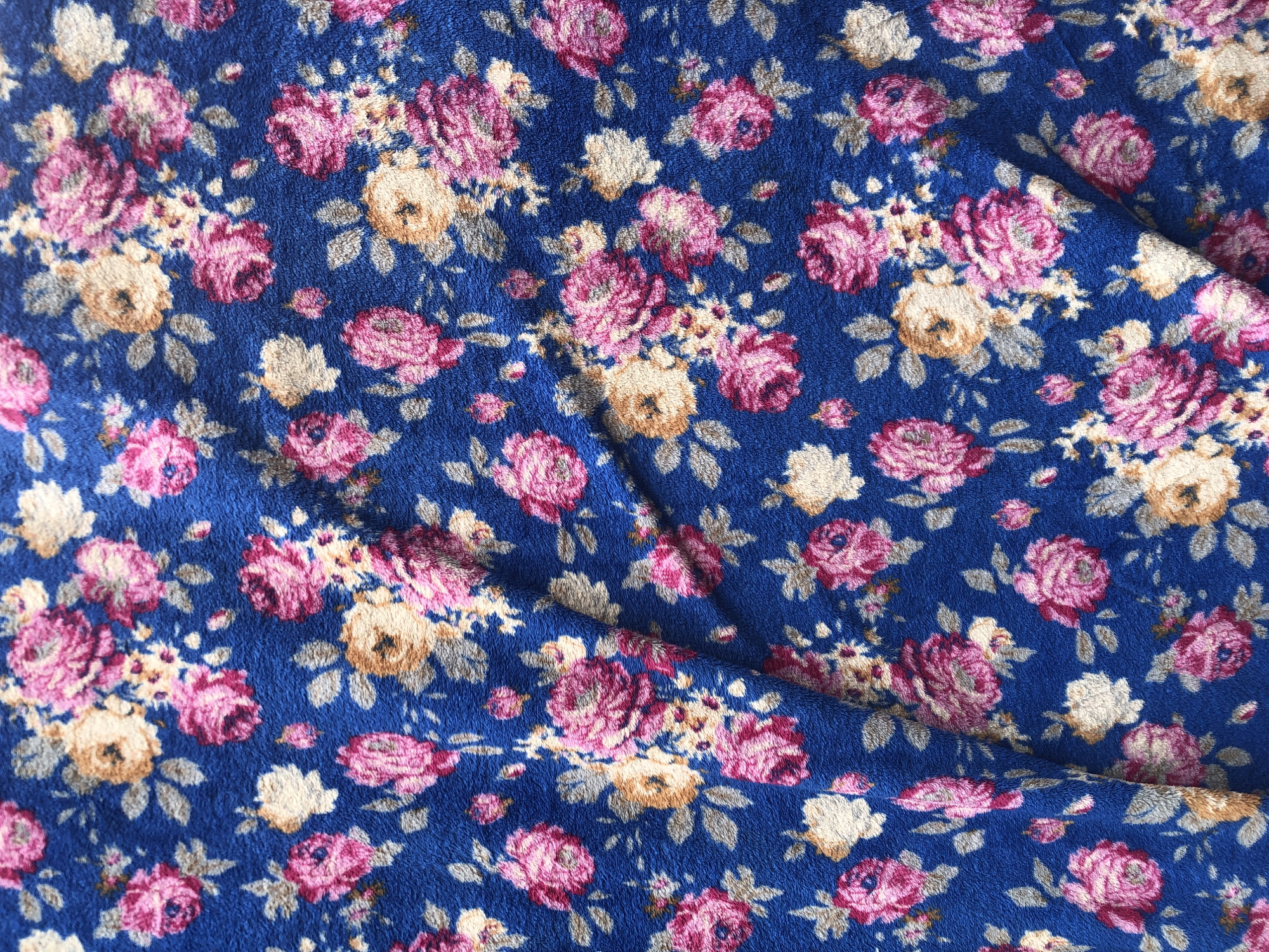 The image is a close-up, overhead view of a blue fabric with a repeating floral pattern. The flowers are rendered in shades of pink, cream, and light brown, with green leaves. The fabric appears to have a soft, slightly textured surface, possibly fleece or a similar material. There are visible folds and creases in the fabric, indicating it is not laid flat. The background is entirely the patterned fabric. There are no people, objects, or environmental features visible outside of the fabric itself. The image does not depict any location in Ado, Nigeria, or any other specific geographic setting.