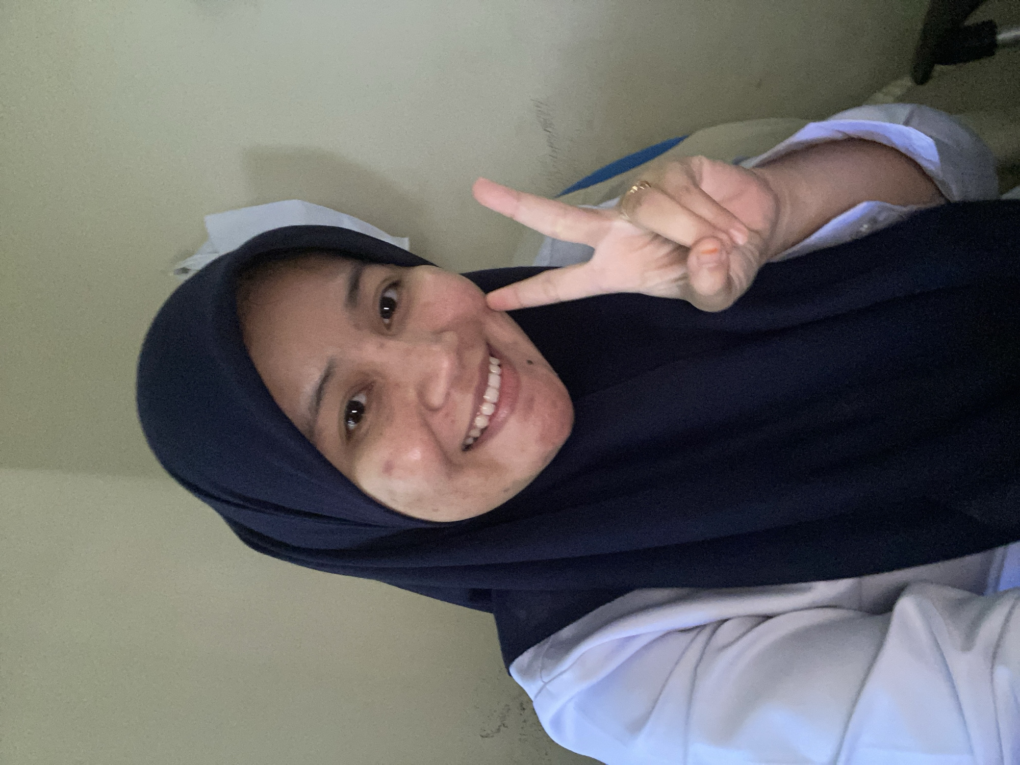 A young woman wearing a dark blue hijab and a white collared shirt is captured in a selfie. She is smiling and making a peace sign with her right hand. The background is a plain, light-colored wall with some subtle texture. The image appears to have been taken indoors, possibly in a residential or office setting, with no identifiable landmarks of Banjarbaru, Indonesia visible.