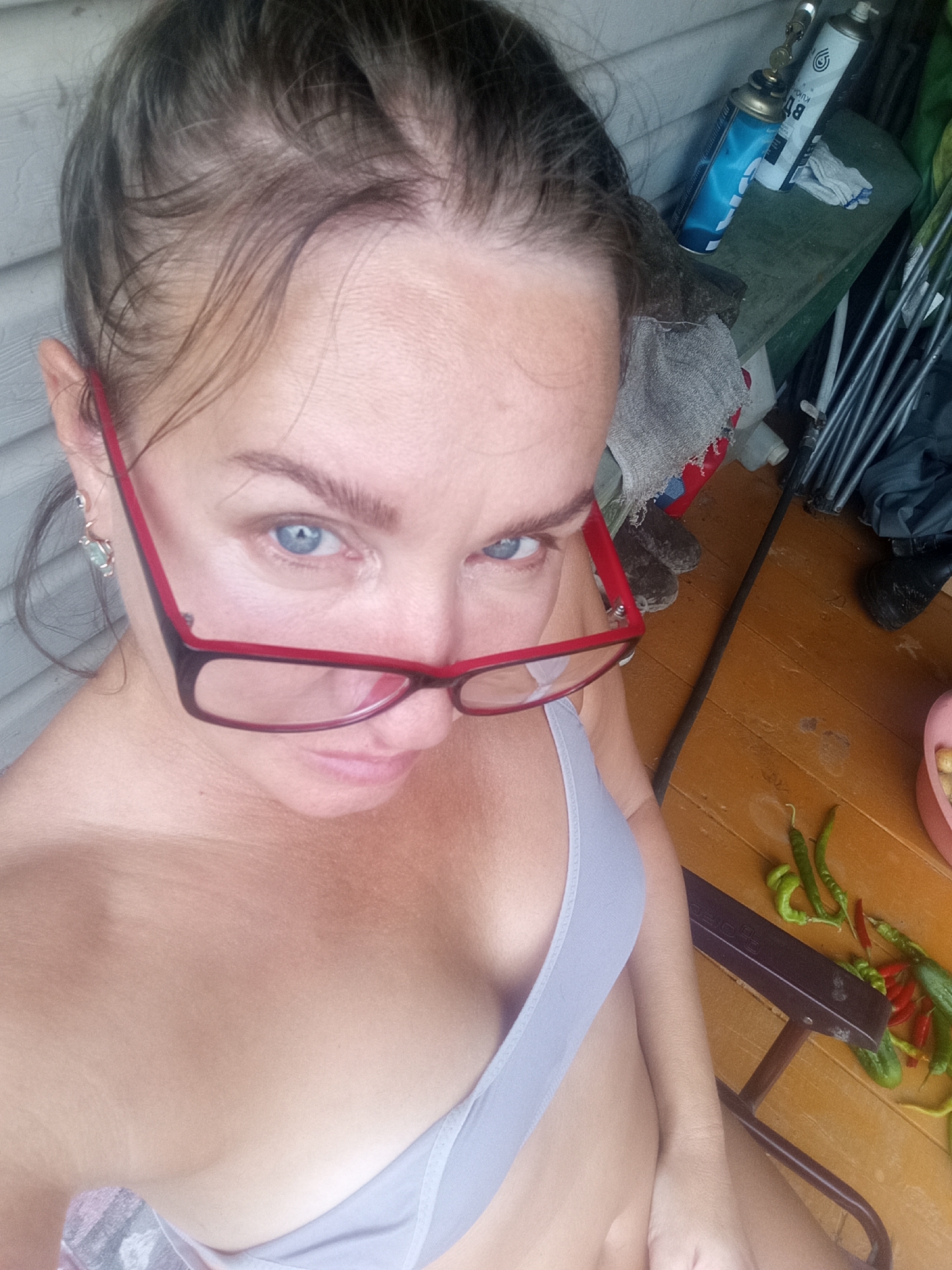 The image is a close-up, high-angle selfie of a woman with light skin and blue eyes, wearing red-rimmed glasses. She is looking directly at the camera with a neutral expression. Her hair is dark brown and pulled back. She is wearing a light blue bra. The background appears to be outdoors, possibly on a wooden porch or deck. Various objects are visible in the background, including a green chair, cans of gas, and some vegetables such as chilies and cucumbers. The overall scene is informal and candid. The location is identified as Novopokrovka, Russia.