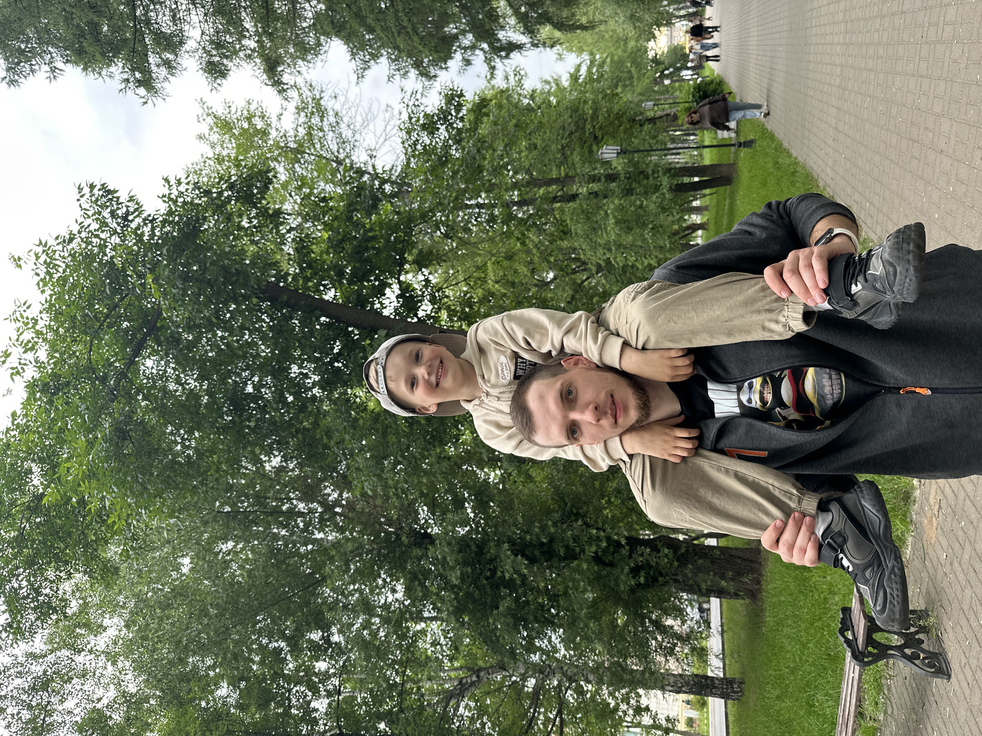A man is carrying a young boy on his shoulders in a park. The man is wearing a dark grey hooded jacket with an orange zipper pull and a t-shirt with a graphic of Teenage Mutant Ninja Turtles. He has short brown hair and a beard, and is looking towards the camera. The boy is wearing a light beige hoodie and light brown pants, and a grey baseball cap. He is smiling and looking towards the camera. They are walking on a paved path lined with trees. In the background, other people are walking on the path, and there are park benches and grass. The image appears to have been taken in the city of Chaikovsky, Russia.