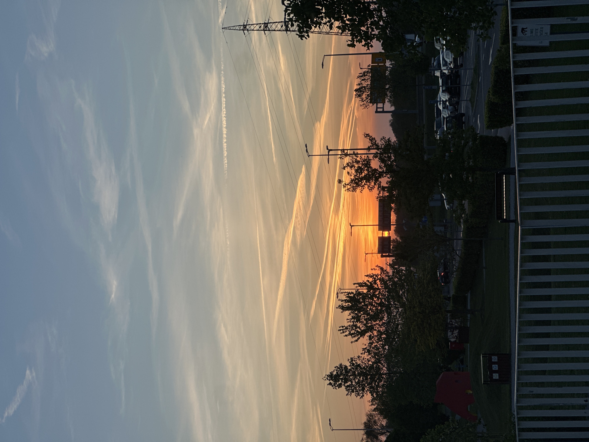 The image shows a sunset scene in Zagreb, Croatia. The sky is filled with streaks of sunlight and contrails from airplanes. There is a large power line tower on the left side of the image. In the middle of the image, there are several street lamps and trees silhouetted against the setting sun. On the right side, there is a parking lot with cars and a building with vertical stripes. In the foreground, there is a playground with a red play structure.