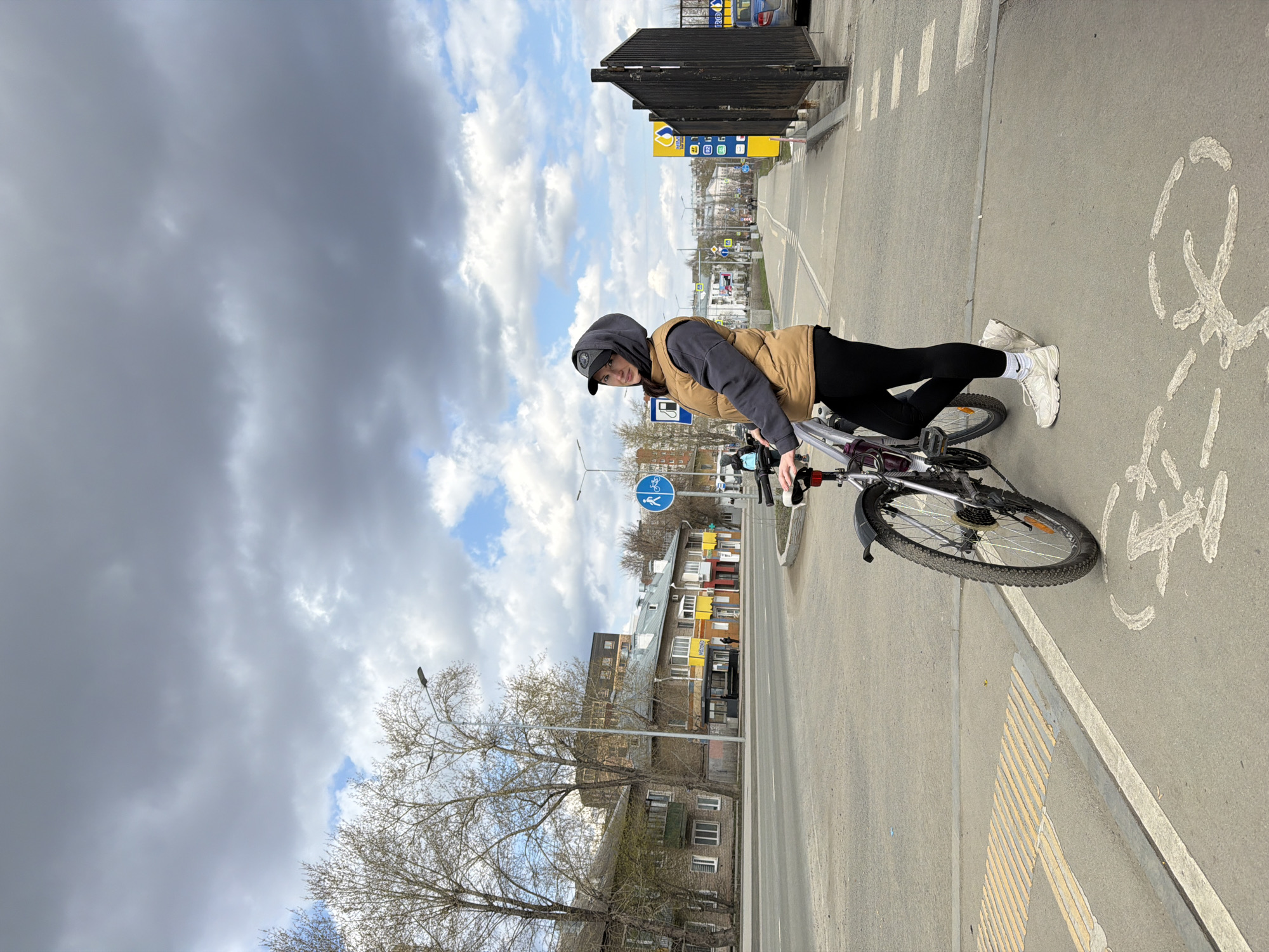 A photograph, rotated 90 degrees counter-clockwise, captures a young woman standing next to her bicycle on a paved path in an urban setting, likely in Perm, Russia. The sky above is largely overcast with large grey clouds, though patches of blue sky and brighter white clouds are visible.
The subject is a young woman with dark hair, wearing a black baseball cap, a dark grey hooded sweatshirt, a light brown puffer vest, black leggings, and white athletic shoes. She stands casually, looking towards the camera with a neutral expression. She is positioned to the right of a silver/grey bicycle, which features dark tires, a black rear fender, and some red accents on the handlebars.
The paved surface below shows clear road markings, including a white painted bicycle symbol indicating a bike path, and a pattern of yellow textured pavement, possibly a tactile warning surface. A round blue sign with white pictograms of a bicycle and a person walking is visible, signifying a shared path. In the background, a street stretches into the distance, flanked by various multi-story buildings of differing architectural styles. Deciduous trees with bare branches or very early buds line the street, suggesting a late winter or early spring season. A dark metal fence or barrier stands near the top right of the frame, and a yellow and blue sign, possibly a gas station logo (e.g., Lukoil), is partially visible further down the road, though its text is illegible. The image depicts a casual moment during the day, with the woman paused on her bicycle journey.