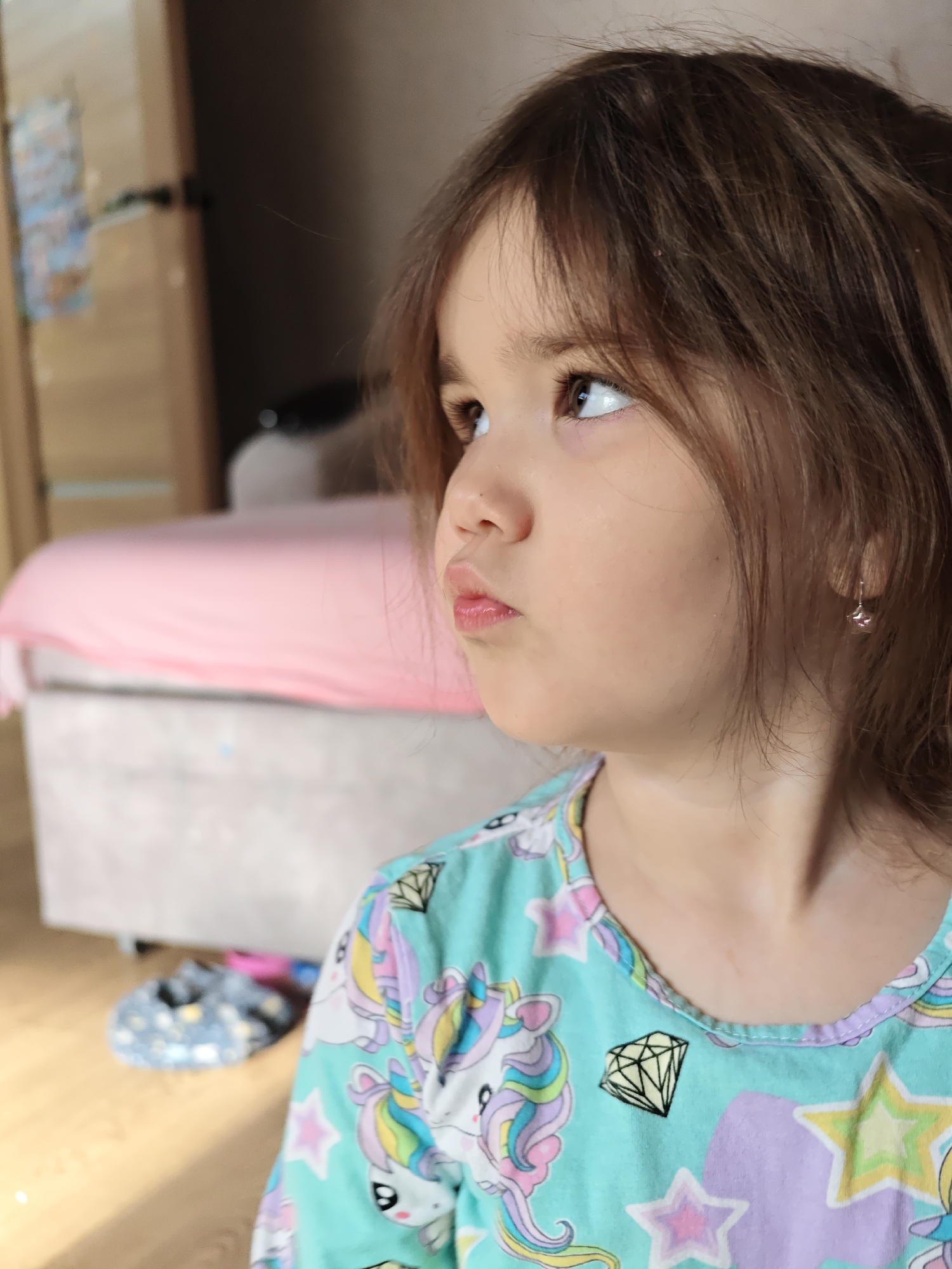 The image captures a close-up, candid moment of a young child, likely a girl, indoors. The child is positioned in the foreground, facing towards the left side of the frame and looking upwards and to the right, showing her left profile.
The child has medium brown hair, which appears slightly unkempt with some loose strands, and bangs that lightly frame her forehead. Her skin tone is light. Her brown eyes are clearly visible, gazing upwards. Her nose is small, and her lips are pursed in a slight pout, suggesting concentration, thoughtfulness, or perhaps a moment of mild contemplation. A small, silver-colored earring, possibly a stud or a small hoop with a charm, is visible in her left earlobe.
She is wearing a light teal or aqua-colored long-sleeved top, possibly pajamas, with a round neckline edged in light purple. The fabric is patterned with cheerful cartoon designs, including unicorns with rainbow-colored manes and tails, various stars (purple, yellow, pink), and black-outlined yellow diamond shapes.
The background is softly blurred, indicating a shallow depth of field. To the immediate left and behind the child, there is a section of a light brown wooden door or a piece of wooden furniture, partially open, revealing a darker interior or shadow. Behind the child's head and extending to the right, a large piece of furniture, likely a bed or sofa, is visible, covered with a bright pink sheet or blanket. The base of this furniture appears to be upholstered in a light grey or beige fabric. In the lower left corner of the background, a light wooden floor is visible, and on it, a pair of dark blue slip-on shoes with a light pattern (possibly dots or stars) can be seen, along with a blurry pink object next to them.
The lighting appears soft and natural, possibly from a window, illuminating the child's face gently. The overall impression is one of a quiet, reflective moment in a relaxed home setting.