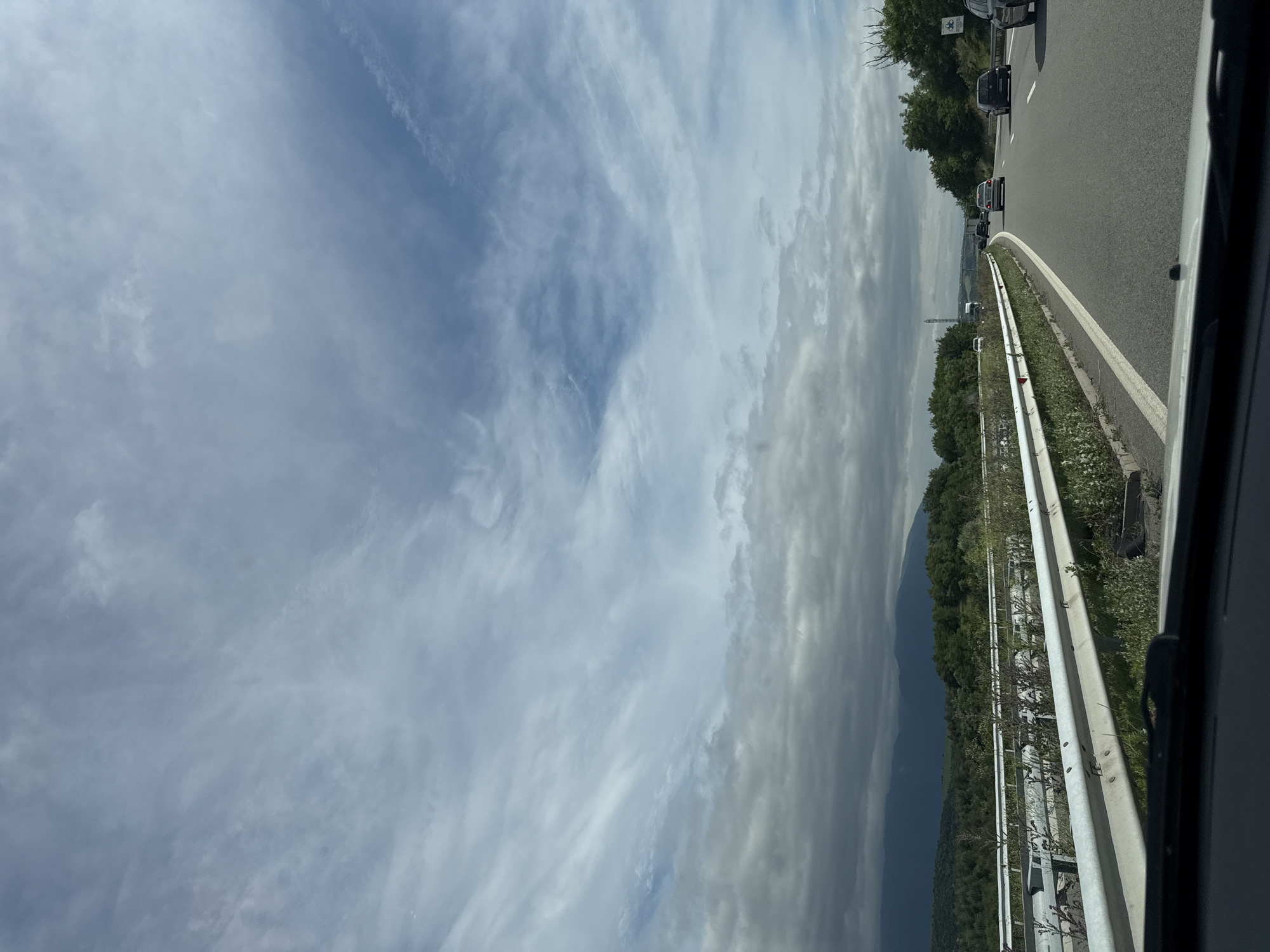 The image shows a highway in the vicinity of Dyakovo, Bulgaria. The perspective is from inside a vehicle, looking forward. The road is a multi-lane highway with a white line marking the lanes and a metal guardrail on the left. Several vehicles are visible on the road ahead, including cars and a truck. The surrounding environment consists of green trees and vegetation along the roadside, with hills or mountains in the distance under a cloudy sky. The sky is partly cloudy with patches of blue sky visible.