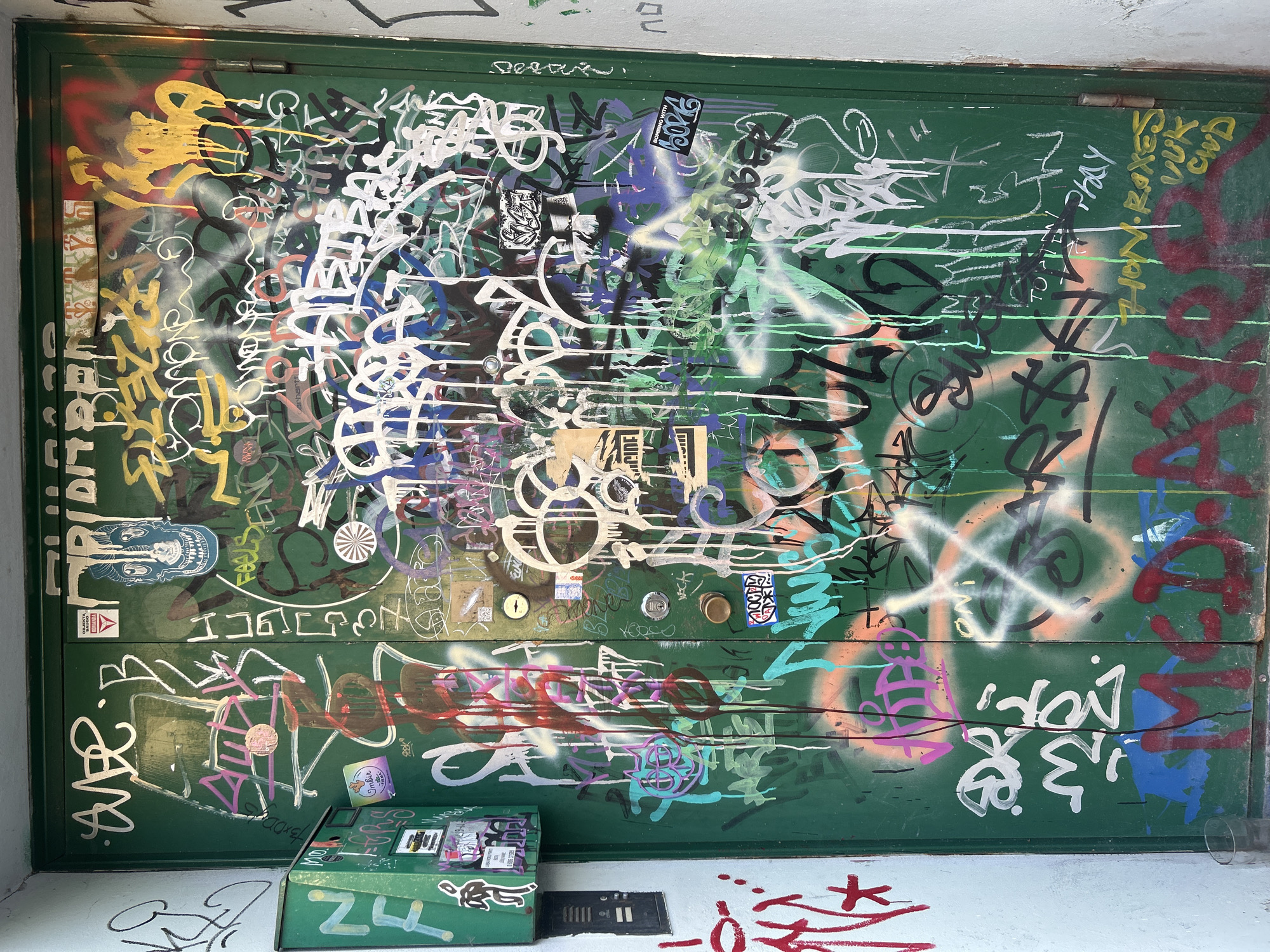 The image depicts a green metal door extensively covered in graffiti. The graffiti is layered and varied in color and style, including tags, lettering, and some cartoonish drawings, such as a blue skull-like face. The door appears to be an exterior door, possibly to a building or an entryway, given the presence of a mailbox or intercom system on the left side of the door. The overall scene is urban and somewhat neglected, suggesting an area with a strong graffiti art presence or urban decay. The door is located in Vilnius, Lithuania, specifically at Vokiečių gatvė 6.