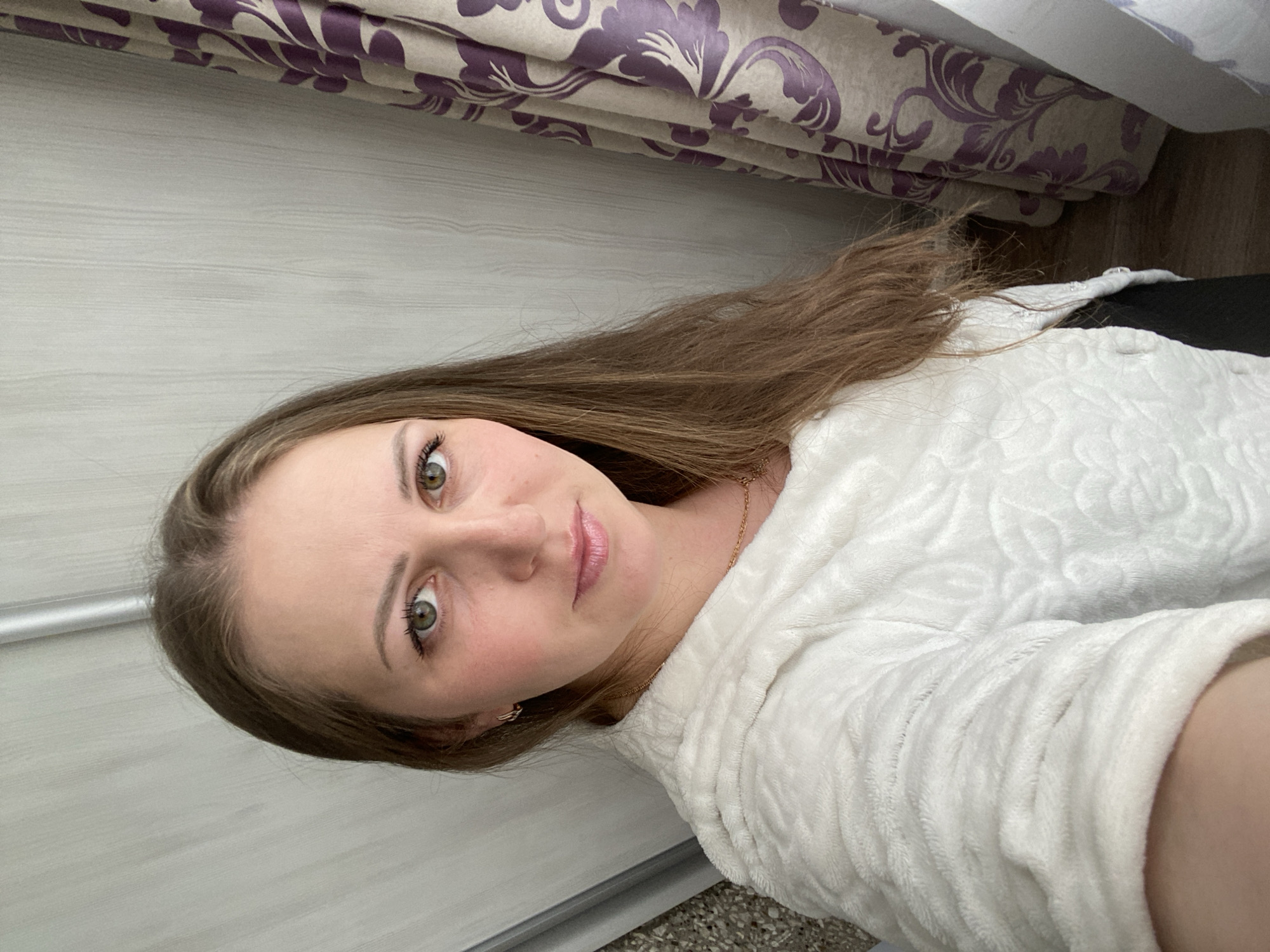 The image shows a woman with long brown hair, wearing a white robe and a gold necklace. She is looking at the camera with a slight smile. The background includes a wall, a curtain with a floral pattern, and a glimpse of the floor. The location is Марковский, Russia.